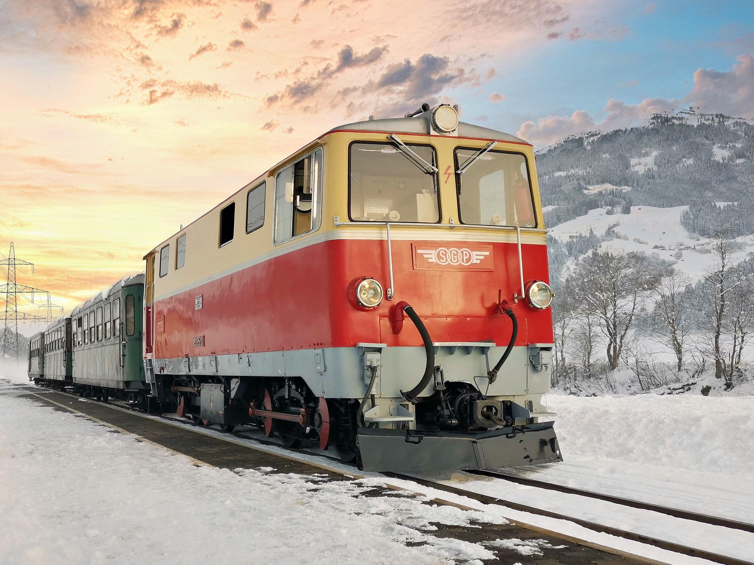 Eine rote Lokomotive steht im Schnee an einem Bahnhof. Im Hintergrund ist eine malerische Winterlandschaft mit Bergen und einem bunten Himmel zu sehen.