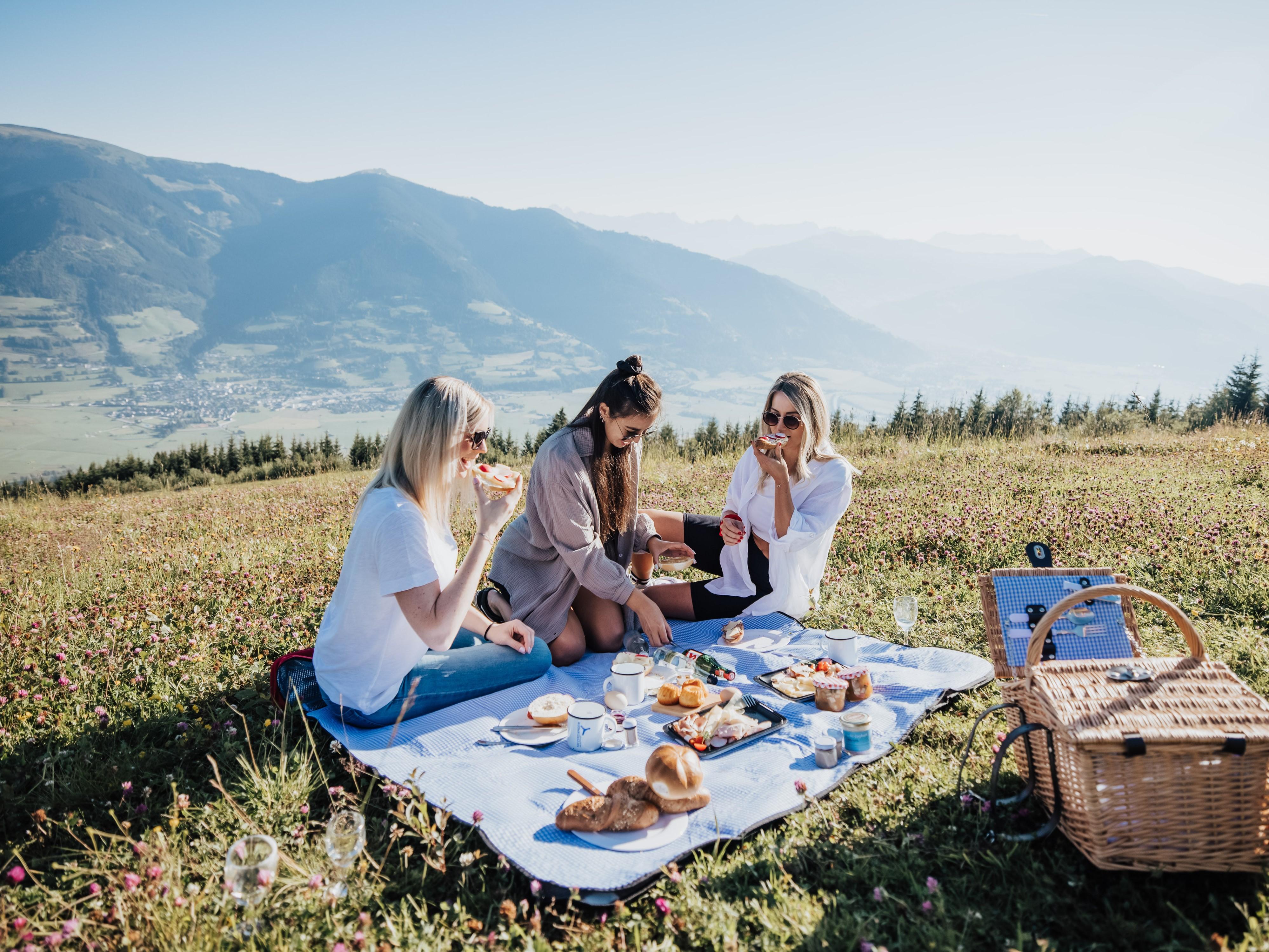Ein Picknick auf einer Wiese in den Bergen. Drei Frauen genießen gemeinsam Essen und Trinken bei sonnigem Wetter.