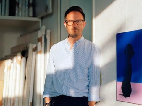 Two musicians are smiling at the camera while sitting next to a piano and a guitar. They are wearing sunglasses and casual clothes.