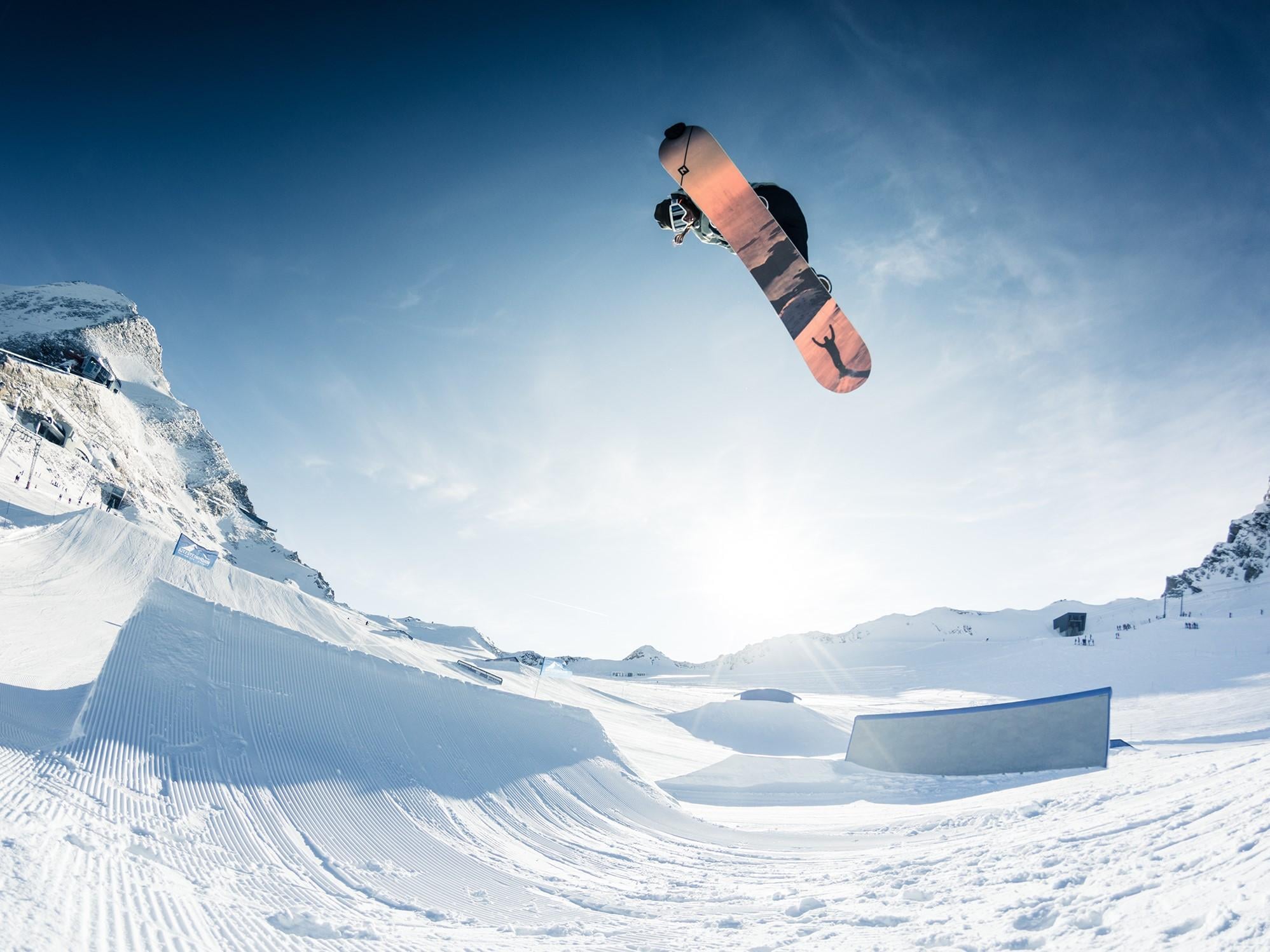 Ein Snowboarder springt über einen Schneepark in den Bergen. Der Himmel ist klar und die Sonne scheint.