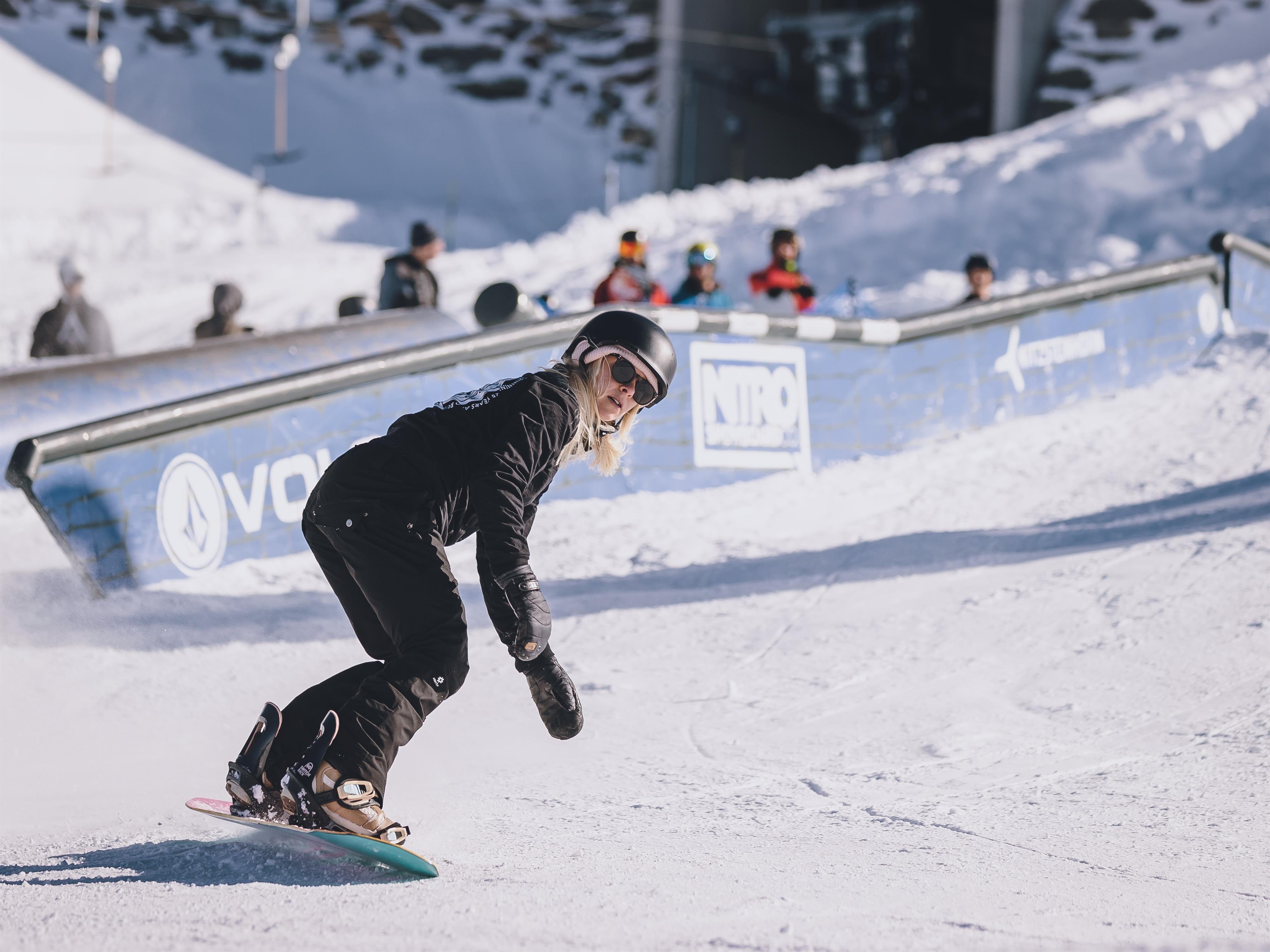 Eine Snowboarderin fährt geschmeidig auf einer schneebedeckten Piste. Im Hintergrund sind Zuschauer und eine Rampe zu sehen.