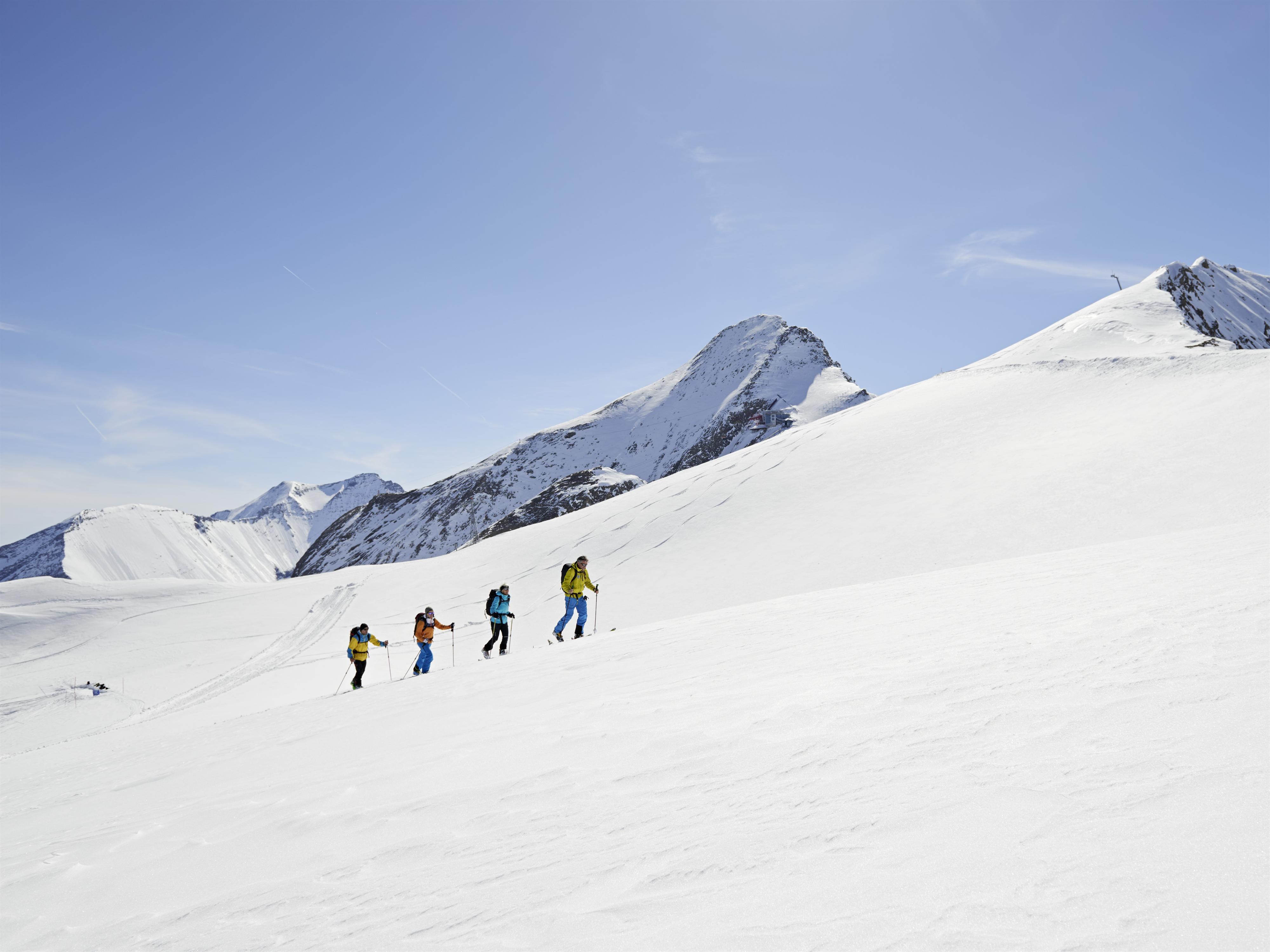 Eine Gruppe von Wanderern geht über eine schneebedeckte Berglandschaft. Der Himmel ist klar und blau, und die Berge sind majestätisch im Hintergrund zu sehen.