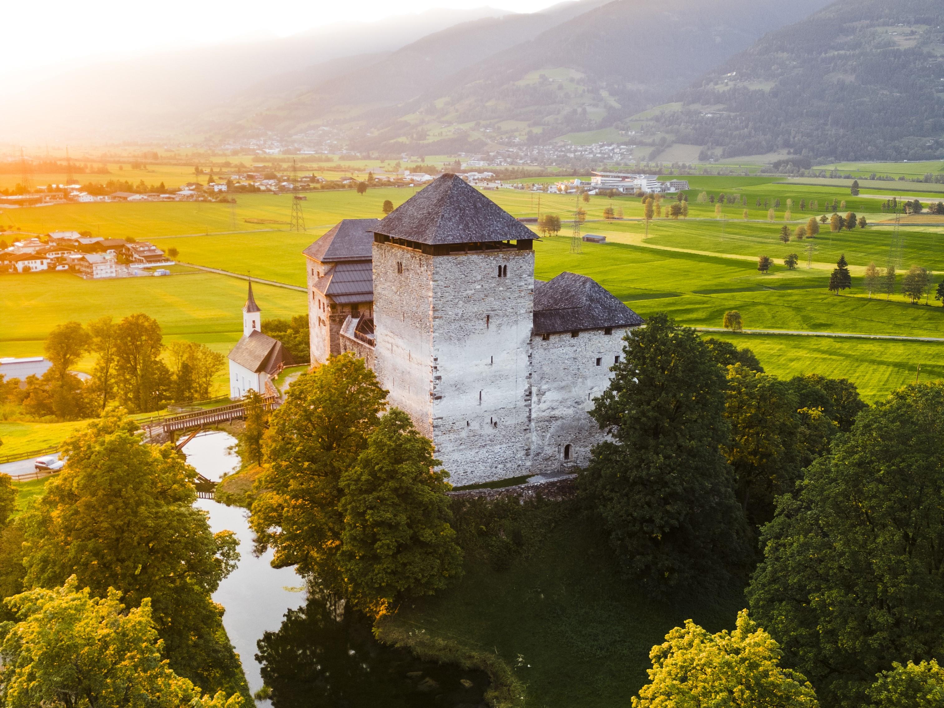 Eine alte Burg umgeben von Grünen Wiesen und Bäumen. Im Hintergrund sind Berge und ein klarer Himmel zu sehen.