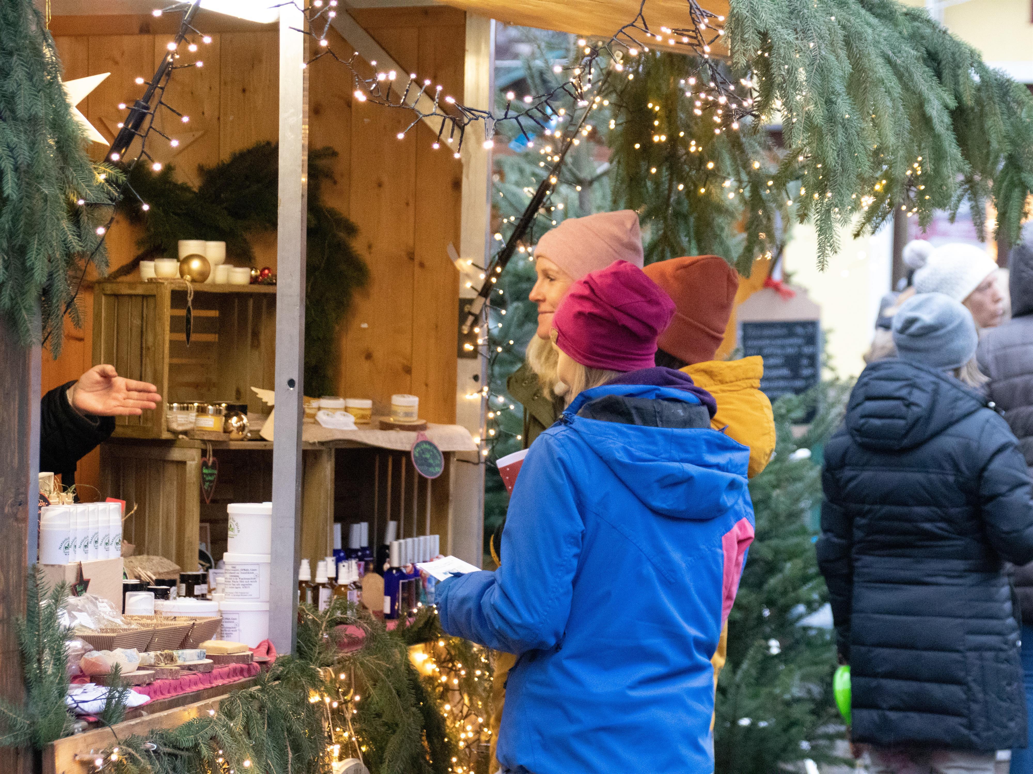 Ein Weihnachtsmarktstand mit festlicher Beleuchtung. Zwei Frauen in bunten Mützen unterhalten sich mit einem Verkäufer.