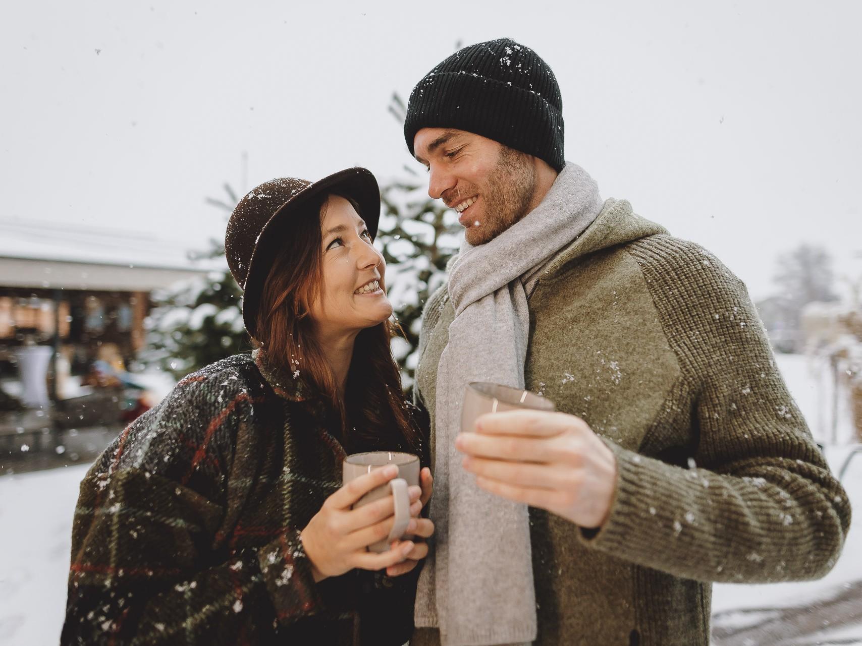 Ein Paar steht in der winterlichen Landschaft und schaut sich liebevoll an. Beide halten Tassen heißen Getränks und sind leicht mit Schnee bedeckt.