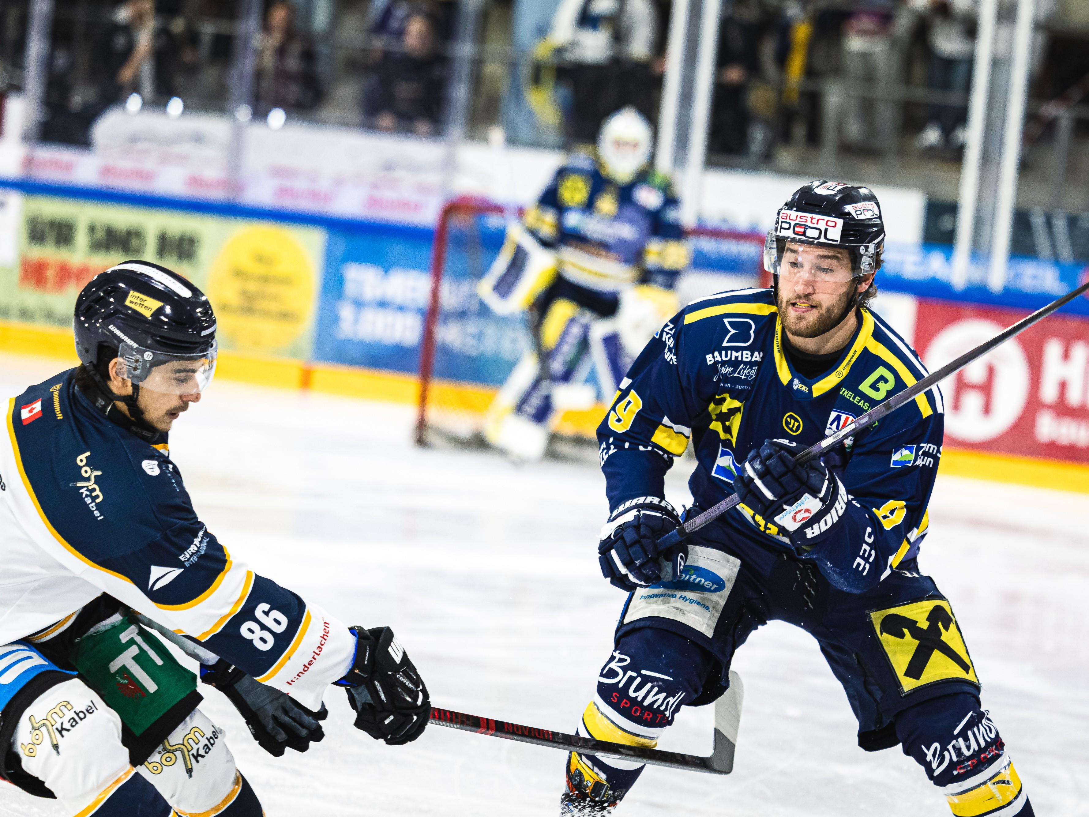 A hockey player in a blue uniform standing on the ice and handling a puck. In the background, spectators and billboards can be seen.