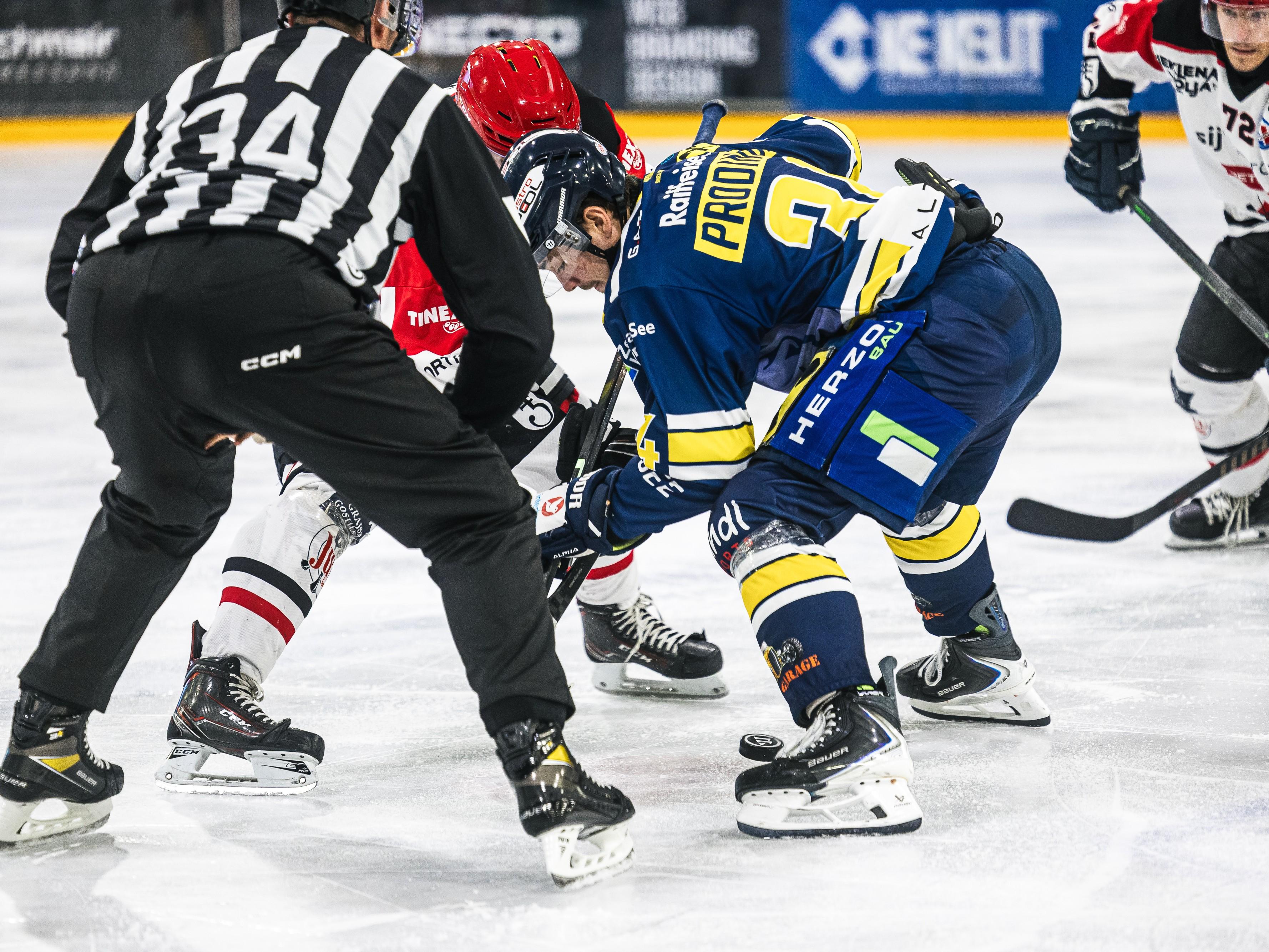 Ein Eishockeyspieler in blauer Uniform, der auf dem Eis steht und einen Puck führt. Im Hintergrund sind Zuschauer und Werbetafeln zu sehen.