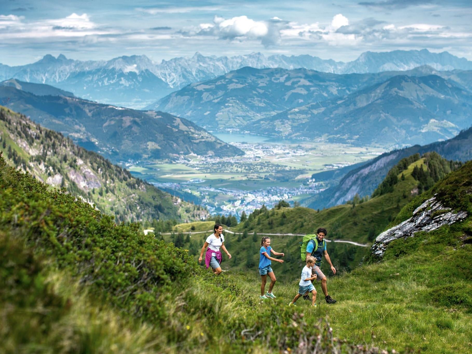 Eine Familie wandert durch eine grüne Berglandschaft. Im Hintergrund sind beeindruckende Berge und ein klarer Himmel zu sehen.