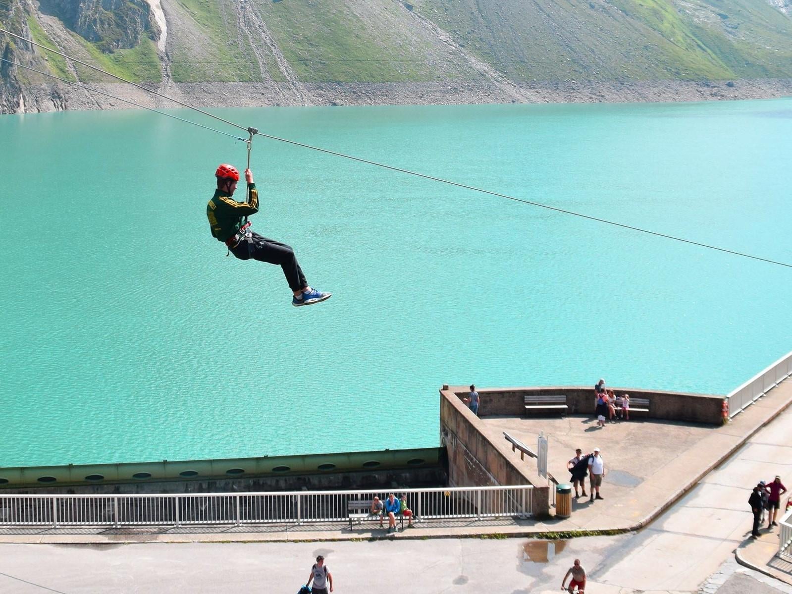 Ein Abenteurer fährt mit der Zipline über einen türkisfarbenen See. Im Hintergrund sind grüne Berge zu sehen.