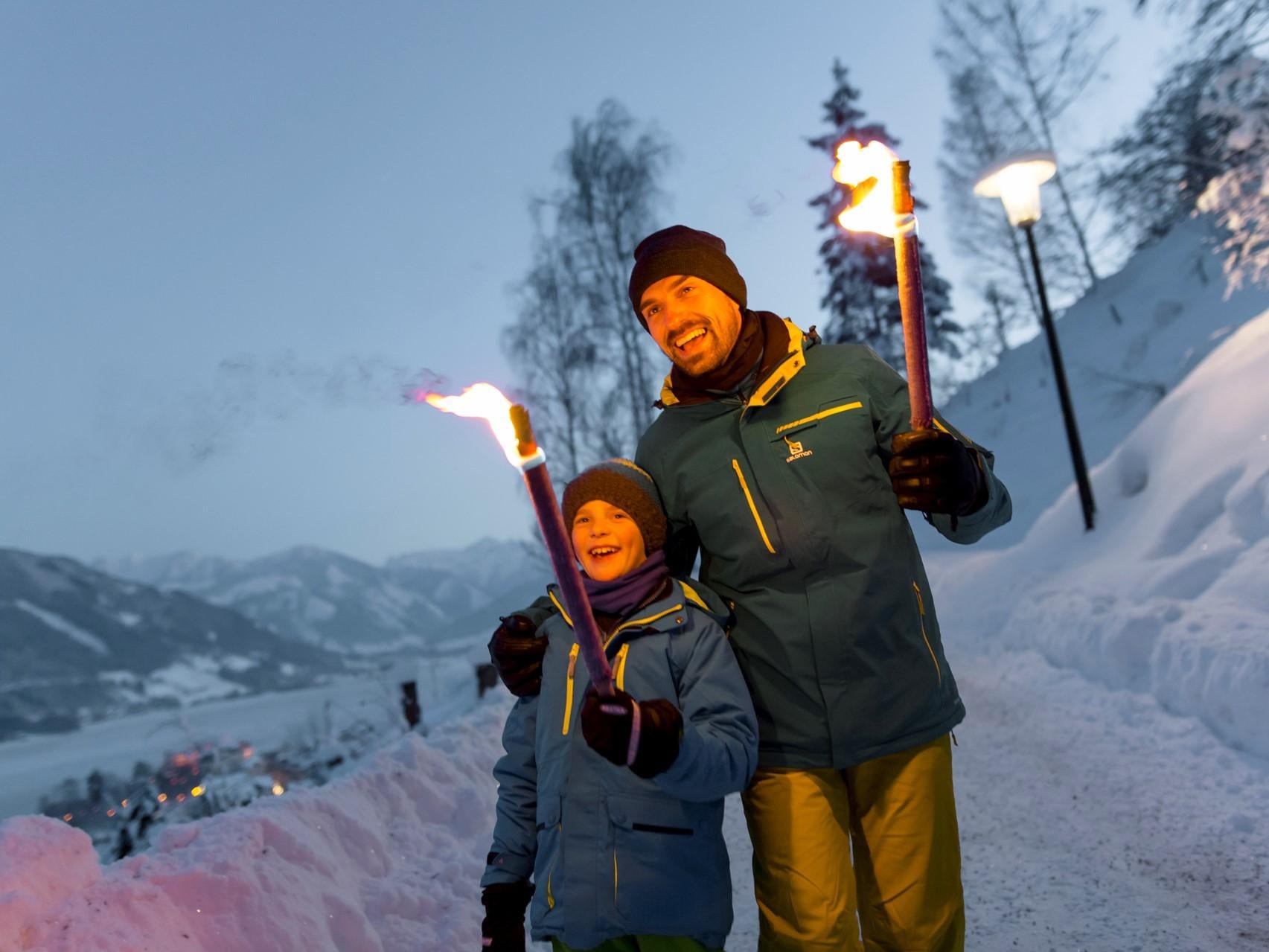 Ein Vater und sein Sohn stehen in einer verschneiten Landschaft und halten Fackeln in den Händen. Der Himmel ist dämmerig und die Umgebung ist mit Schnee bedeckt.