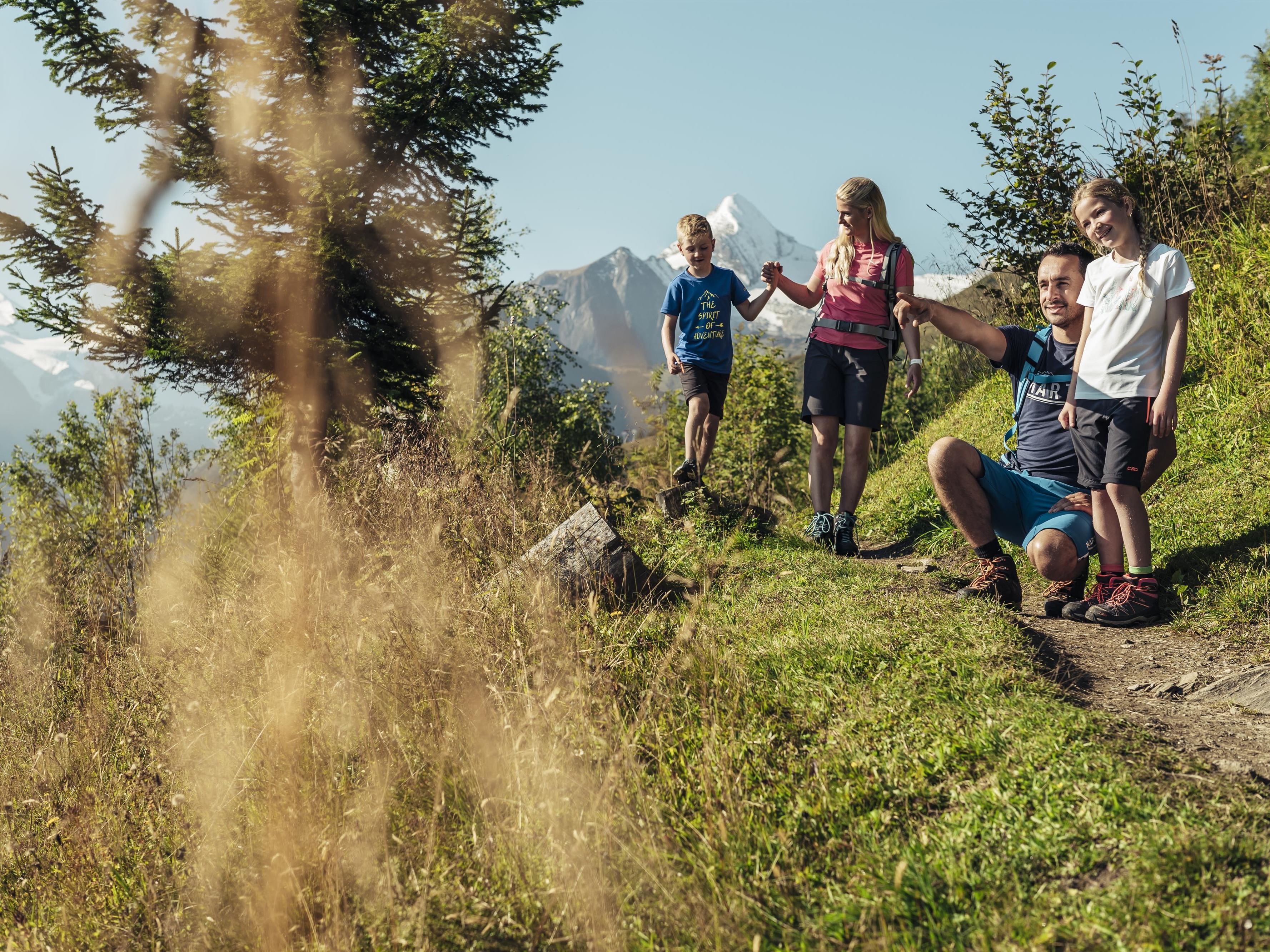 Eine Gruppe von Kindern und einem Erwachsenen wandert auf einem malerischen Weg in den Bergen. Im Hintergrund sind hohe Berge und die Natur zu sehen.