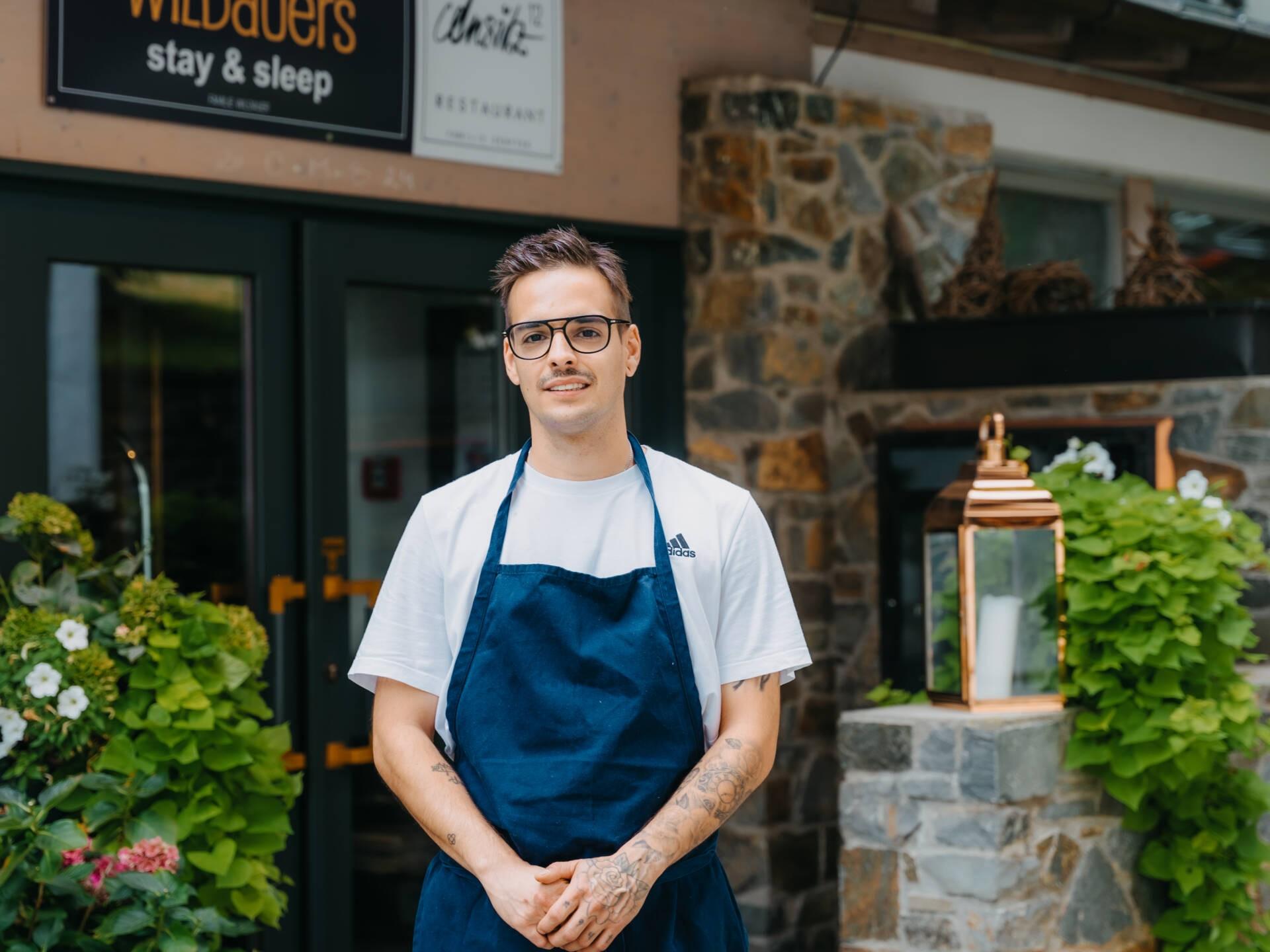 Zwei Köche stehen vor einer Holzwand und machen eine ernsthafte Pose. Sie tragen weiße Kochjacken und wirken konzentriert.