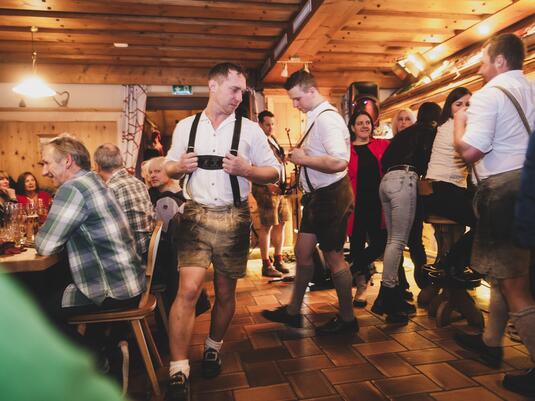 Eine lebhafte Szenerie in einem traditionellen Gasthaus mit Menschen in Trachten. Viele Gäste interagieren und genießen die Atmosphäre.