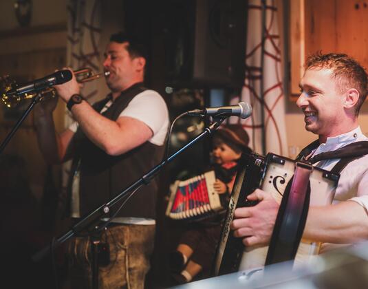 Zwei Musiker spielen Instrumente auf einer Bühne. Im Hintergrund steht ein Puppenspieler.