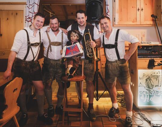A group of four men in traditional Bavarian clothing stands together while a child sits on a chair. They hold musical instruments and smile at the camera.