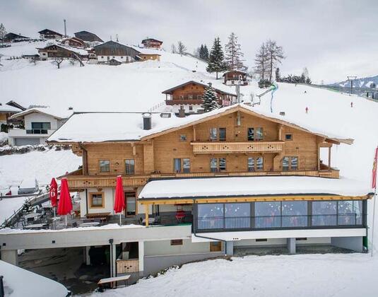 A cozy wooden house in a snowy mountain landscape. In the foreground, red umbrellas and ski lifts are visible.