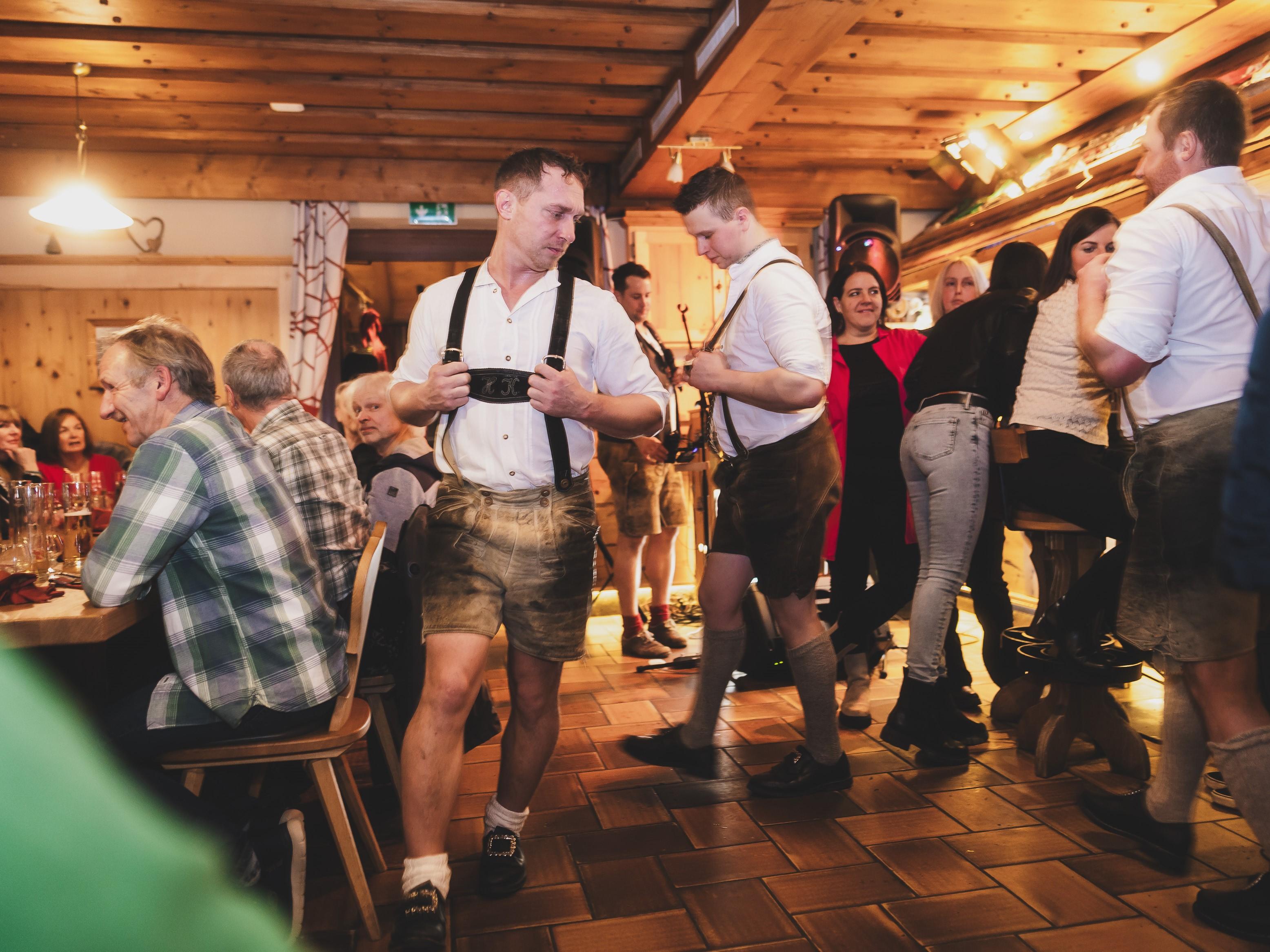 Eine lebhafte Szenerie in einem traditionellen Gasthaus mit Menschen in Trachten. Viele Gäste interagieren und genießen die Atmosphäre.