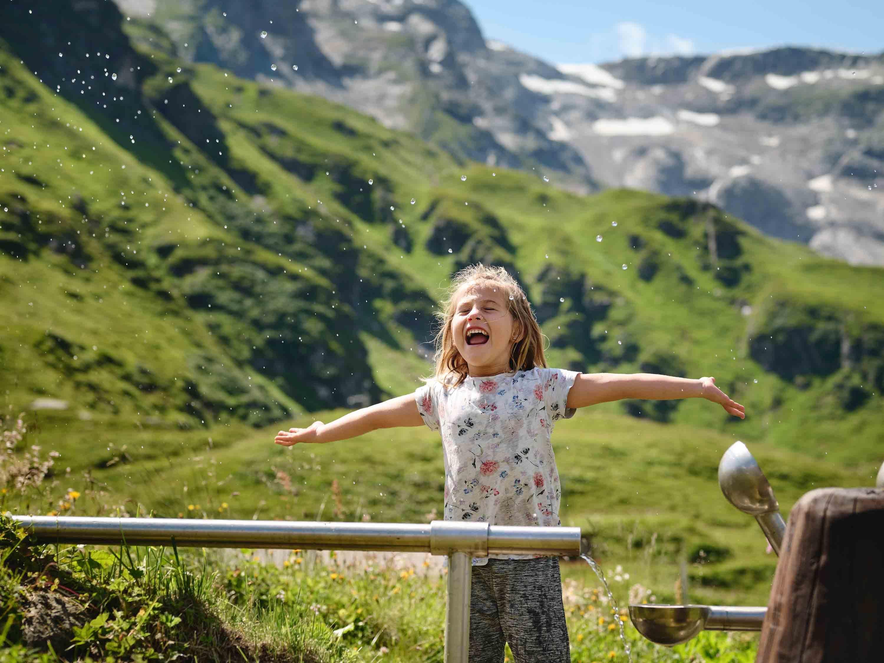 Ein fröhliches Mädchen steht in der Natur und breitet die Arme aus. Im Hintergrund sind grüne Berge und blauer Himmel zu sehen.
