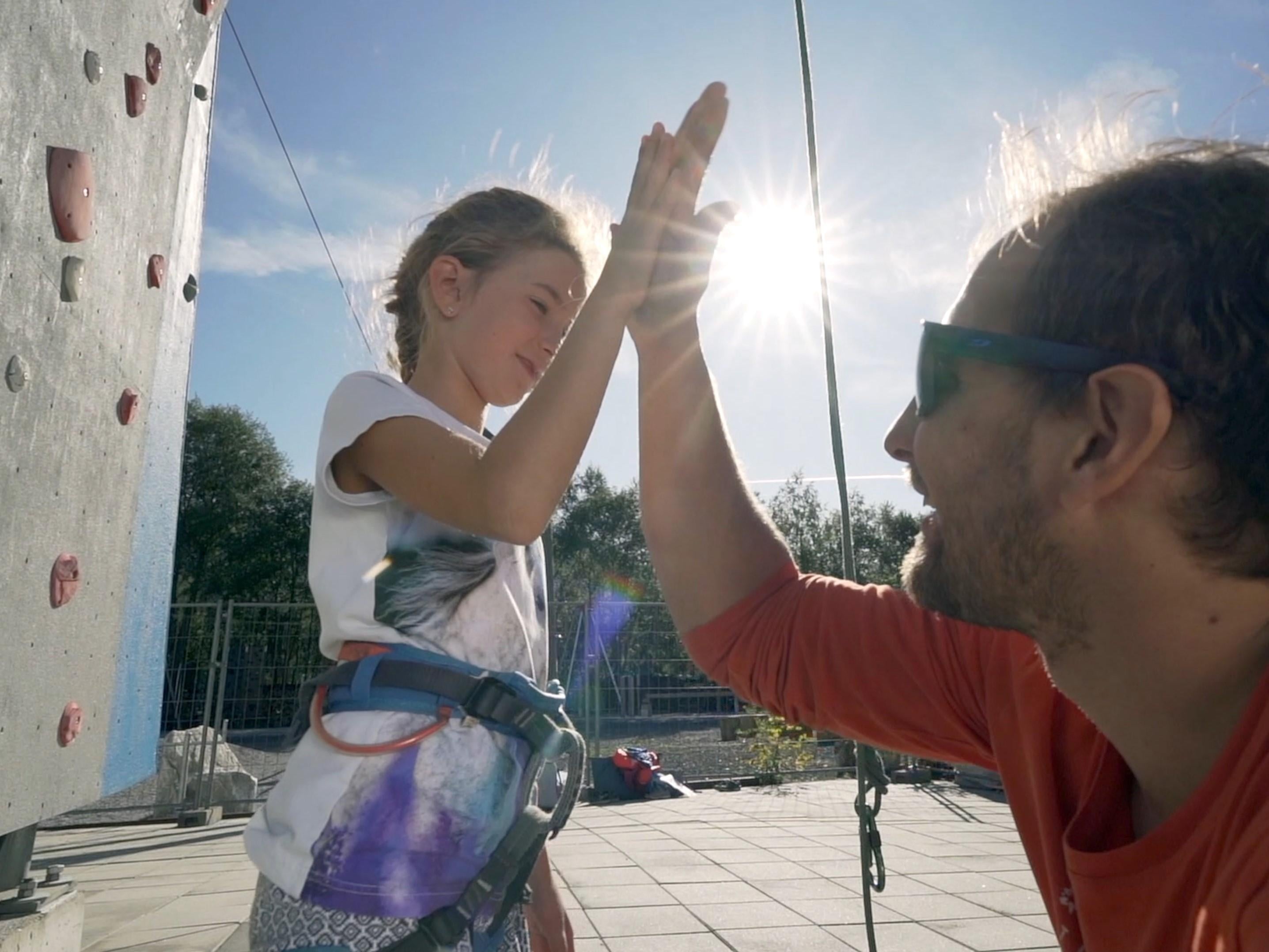 Ein Erwachsener und ein Kind machen ein High-Five vor einer Kletterwand. Die Sonne scheint hell im Hintergrund.