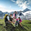 A family is sitting on a meadow in the mountains, enjoying nature. In the background, there is a cable car and snow-covered mountains.