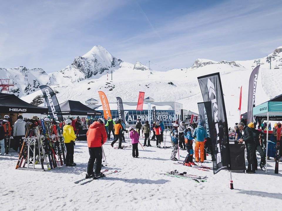 Eine Snowboarderin fährt geschmeidig auf einer schneebedeckten Piste. Im Hintergrund sind Zuschauer und eine Rampe zu sehen.