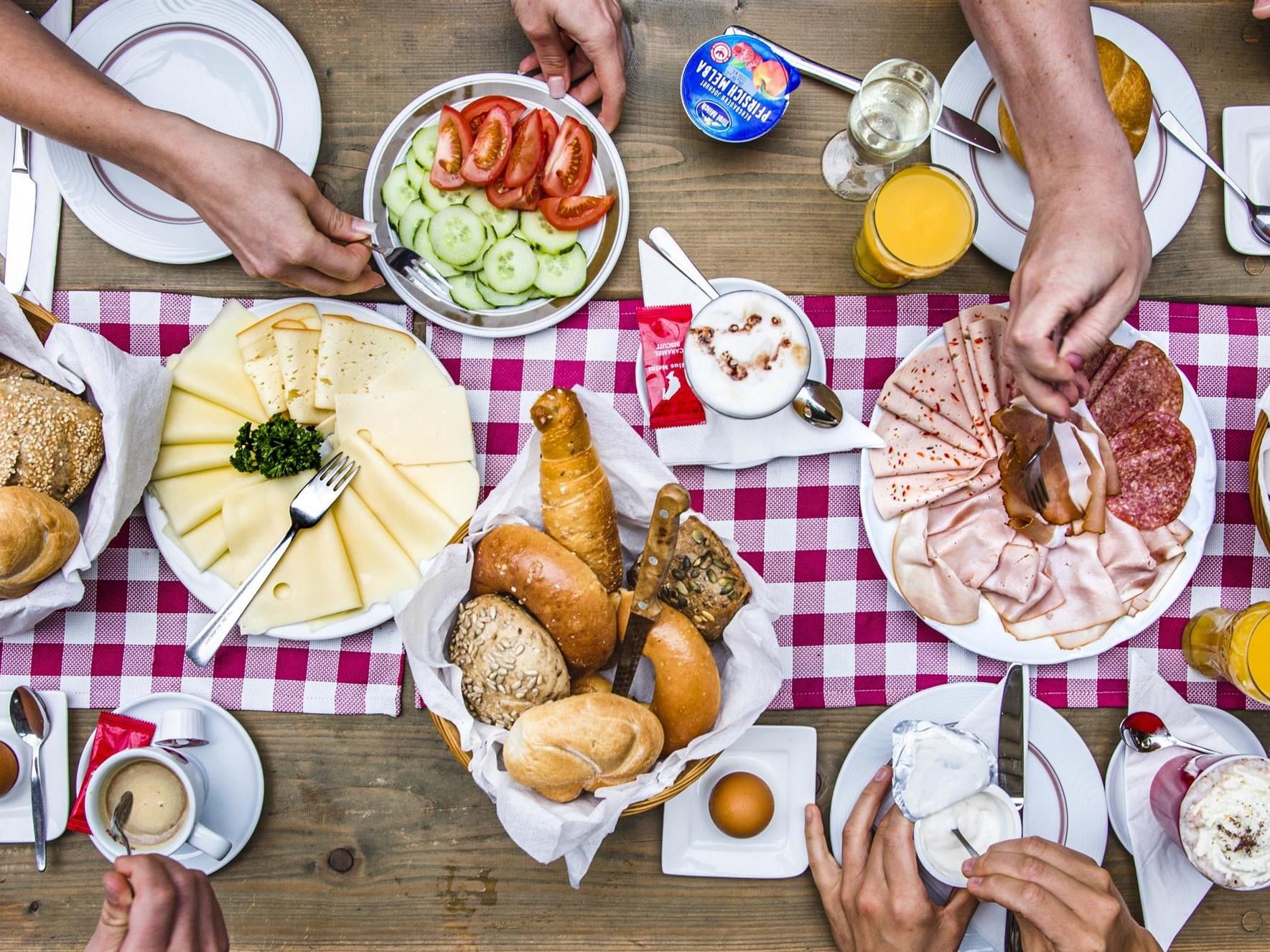 Ein reichhaltiges Frühstück mit verschiedenen Brotsorten, Käse, Wurst, Gemüse und Getränken. Die gedeckte Tischdecke in Karomuster sorgt für eine einladende Atmosphäre.