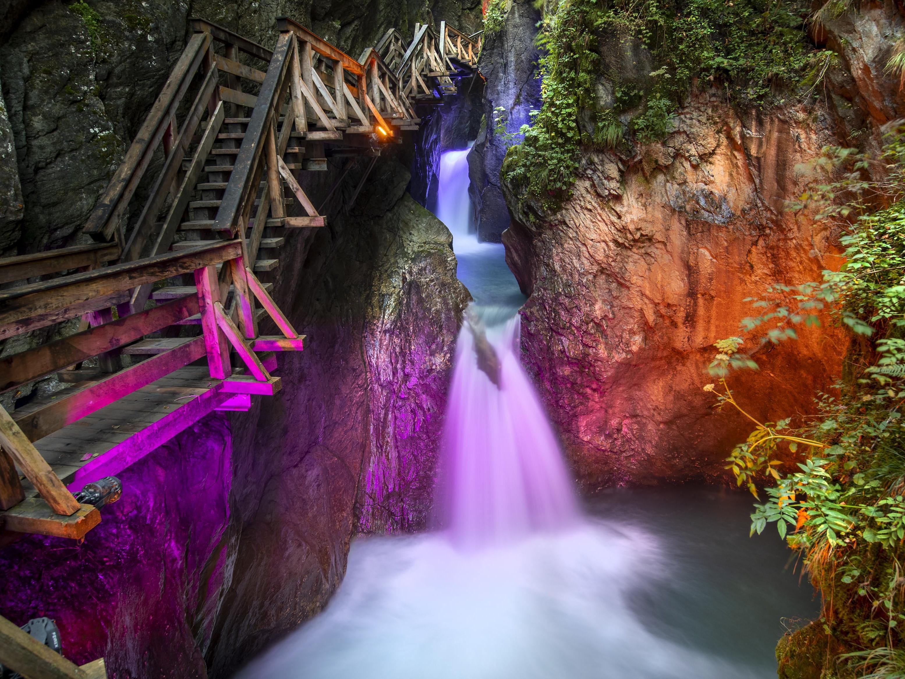 Ein malerischer Wasserfall in einer Schlucht, umgeben von Felsen und üppigem Grün. Ein Holzweg verläuft entlang des Wassers und schafft eine einladende Atmosphäre.