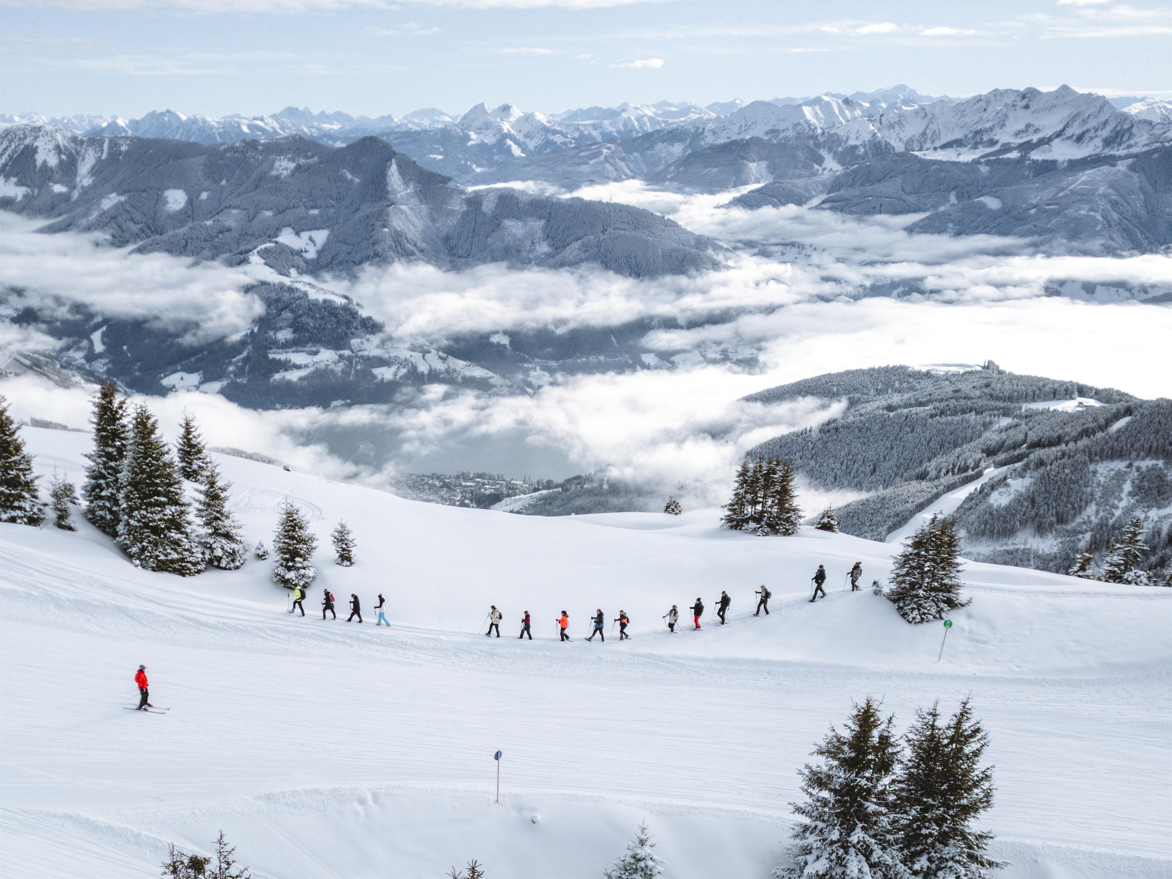 Eine Gruppe von Skifahrern bewegt sich über eine verschneite Landschaft. Im Hintergrund sind Berge und Wolken sichtbar.