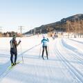 Zwei Langläufer auf einer schneebedeckten Strecke. Im Hintergrund sind Bäume und Berge zu sehen.