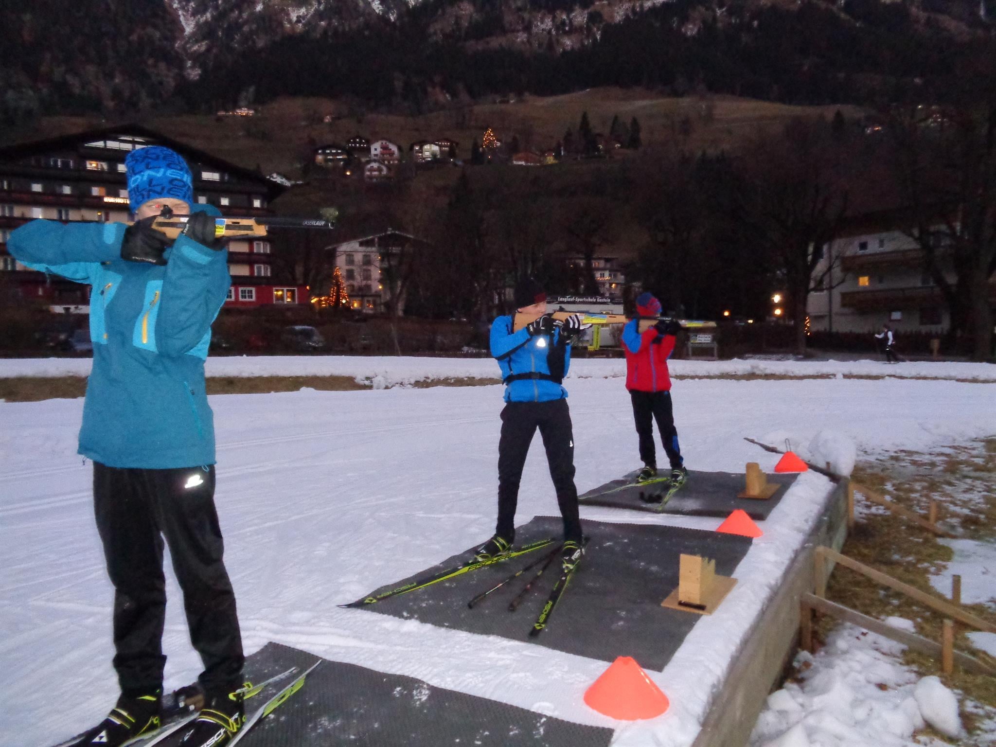 Drei Skilangläufer stehen auf einer Schneebahn und üben das Schießen. Im Hintergrund sind beleuchtete Häuser und Berge zu sehen.