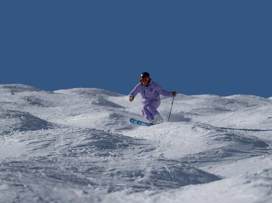 A skier in purple gear is skiing across a snow-covered landscape. The sky is clear and blue.