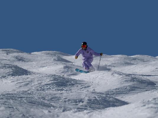 A skier in purple gear is skiing across a snow-covered landscape. The sky is clear and blue.