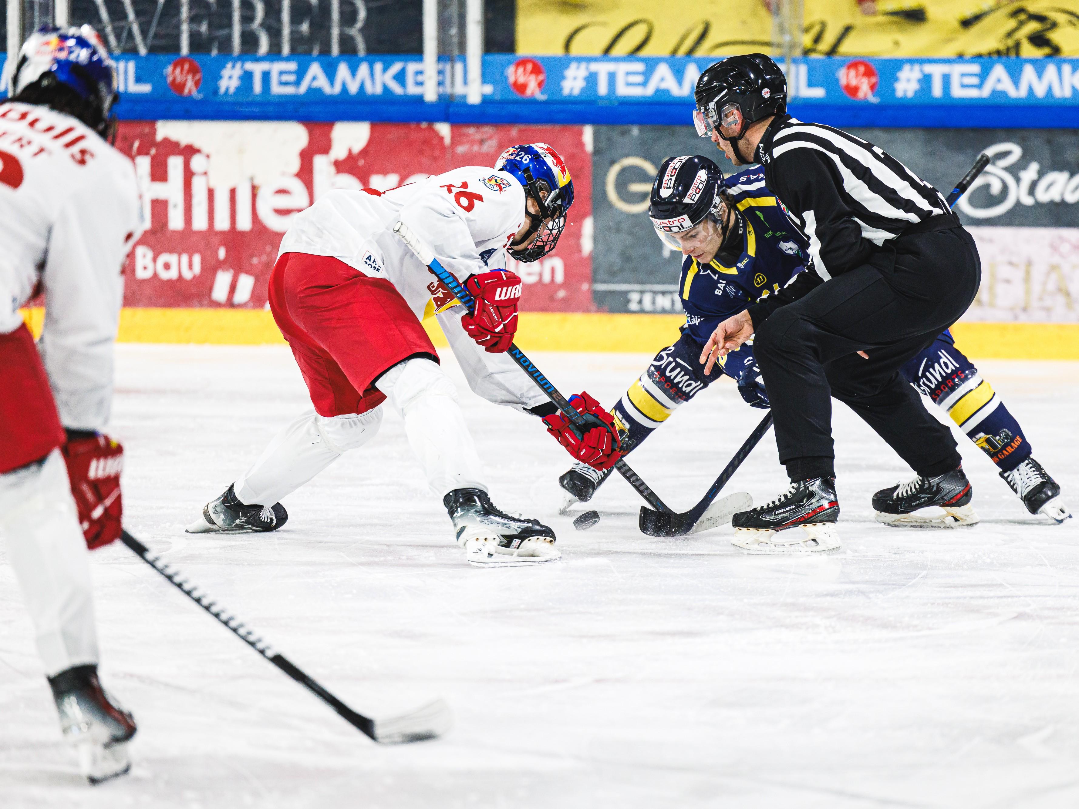 Ein Eishockeyspieler in blauer Uniform, der auf dem Eis steht und einen Puck führt. Im Hintergrund sind Zuschauer und Werbetafeln zu sehen.