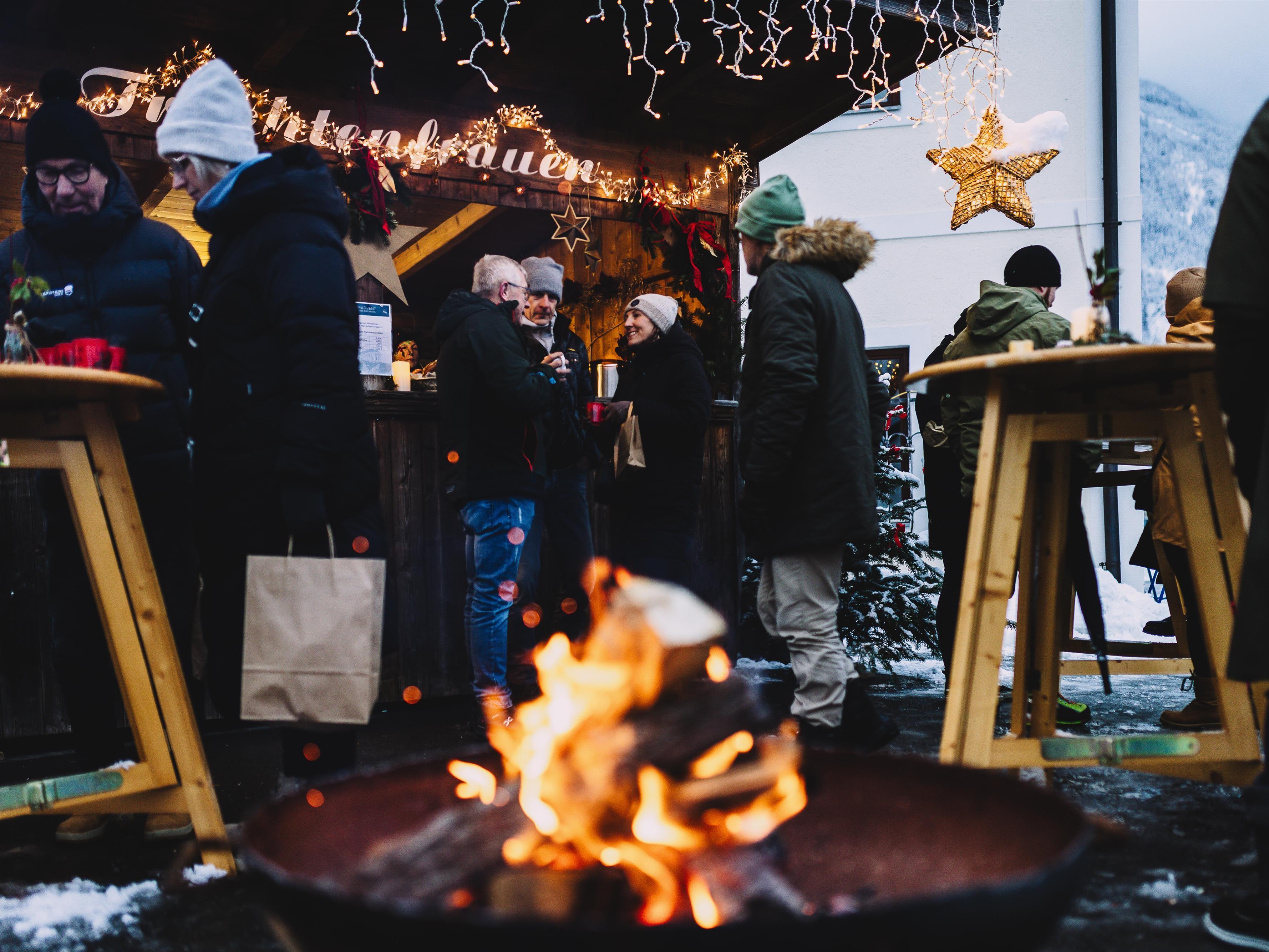 Ein festlicher Weihnachtsmarkt mit einem warmen Feuer im Vordergrund. Menschen stehen in gemütlicher Atmosphäre zusammen und genießen die Stimmung.