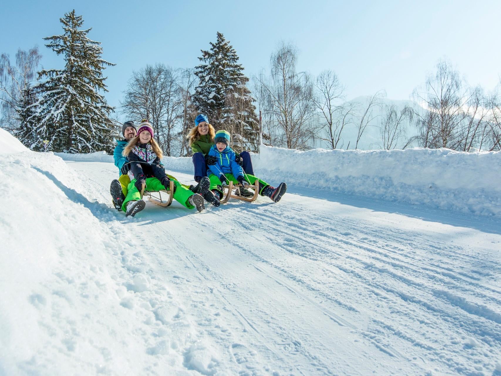 Eine Gruppe von Freunden rutscht fröhlich einen verschneiten Hang hinunter. Die Sonne scheint und die Bäume sind mit Schnee bedeckt.
