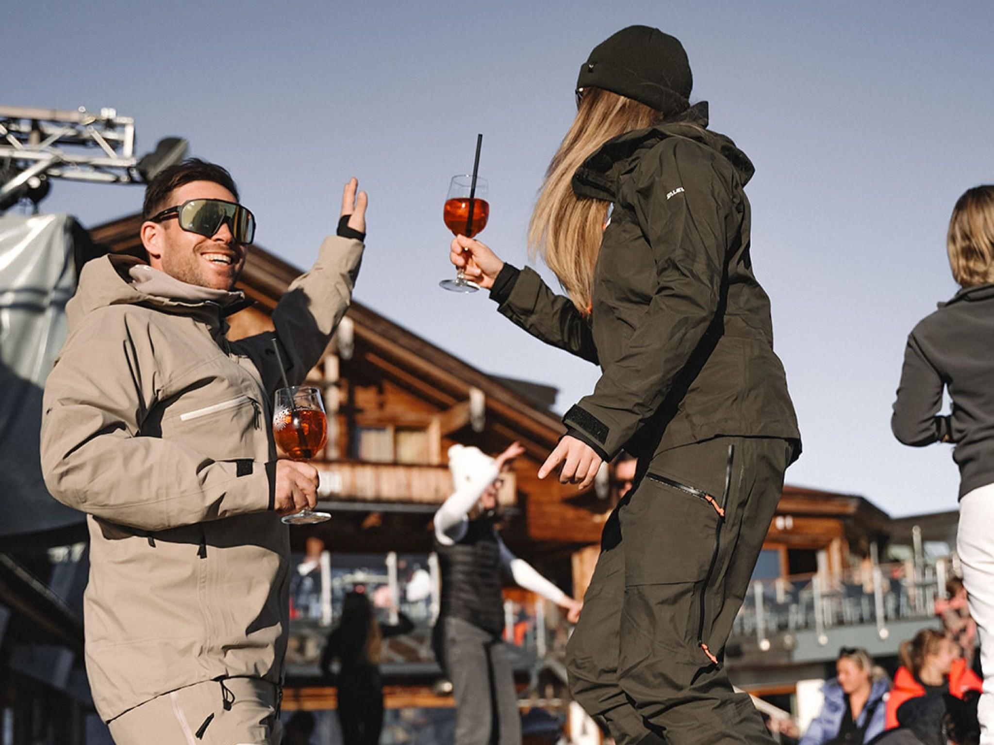 Zwei Personen tanzen fröhlich in winterlicher Kleidung auf einer Terrasse. Im Hintergrund sind Holzgebäude und weitere Gäste zu sehen.