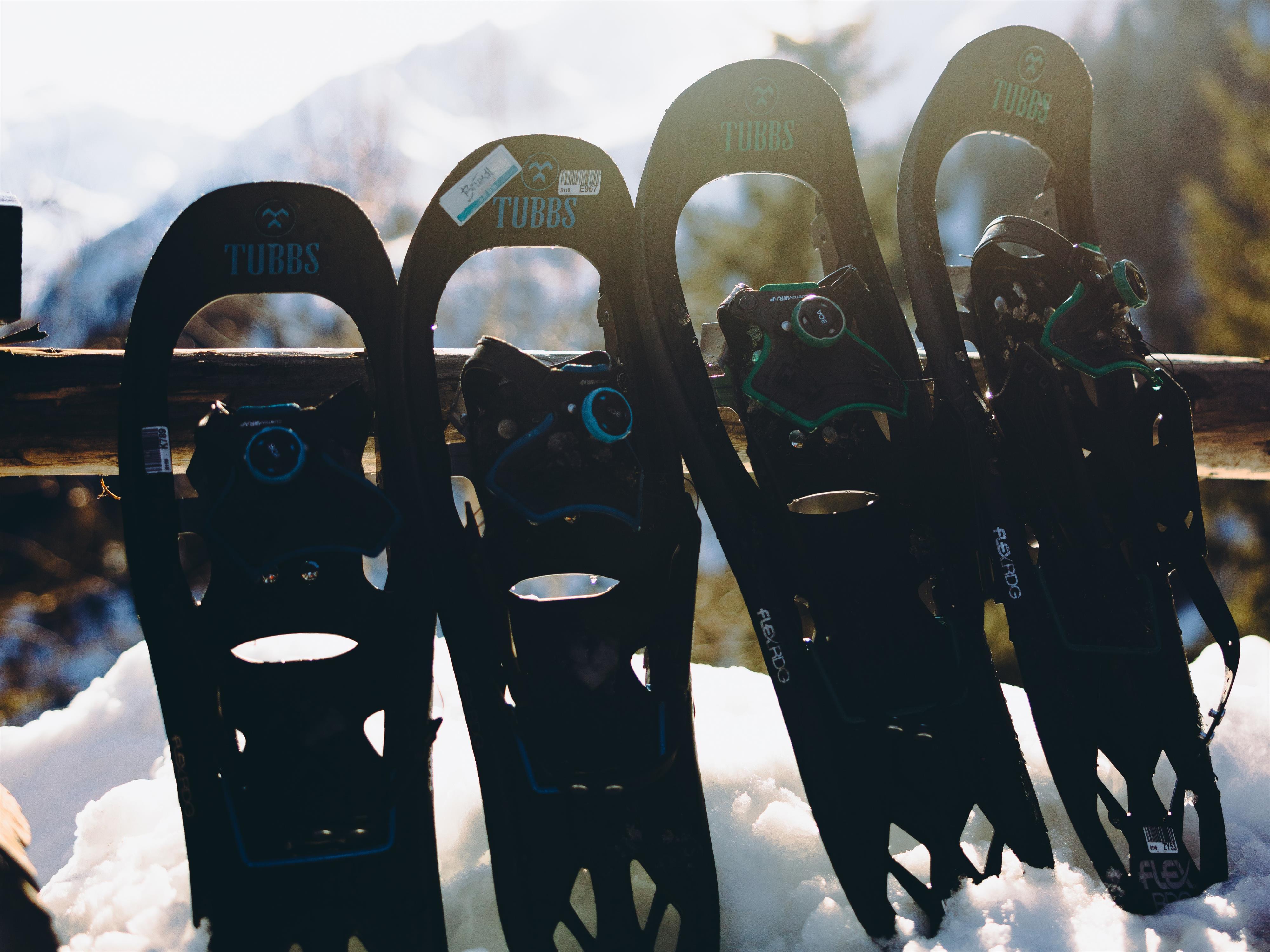 Vier Schneeschuhe stehen auf einem Holzgeländer im Schnee. Im Hintergrund sind die schneebedeckten Berge und ein klarer Himmel zu sehen.