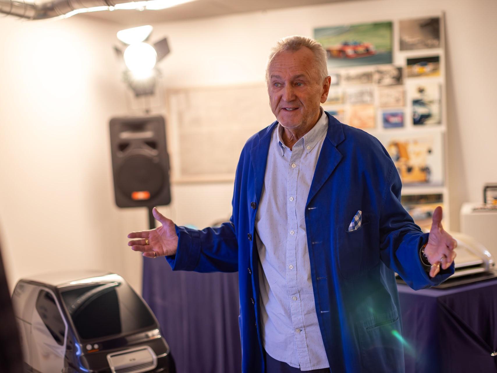 Ein älterer Mann in blauer Arbeitskleidung spricht vor Publikum. Im Hintergrund sind Auto-Modelle und Bilder zu sehen.