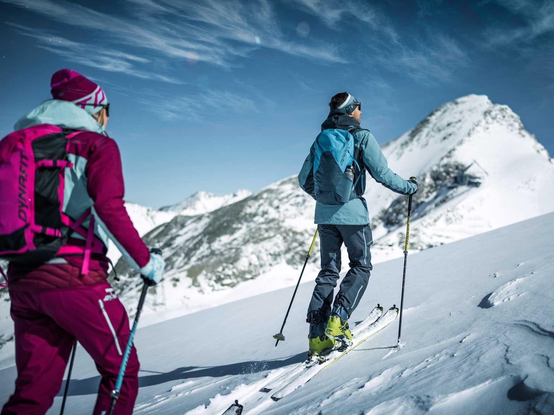 Zwei Skitourengeherinnen wandern durch verschneite Berge. Der Himmel ist klar und die Landschaft ist beeindruckend.