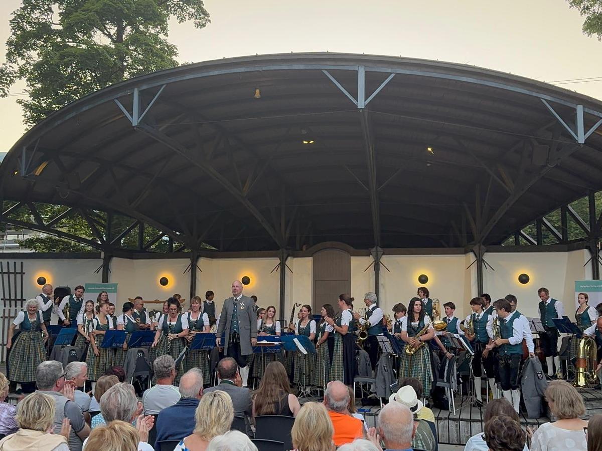 Ein Musikensemble führt ein Konzert unter einem schattigen Pavillon auf. Die Zuschauer genießen die Darbietung im Freien.