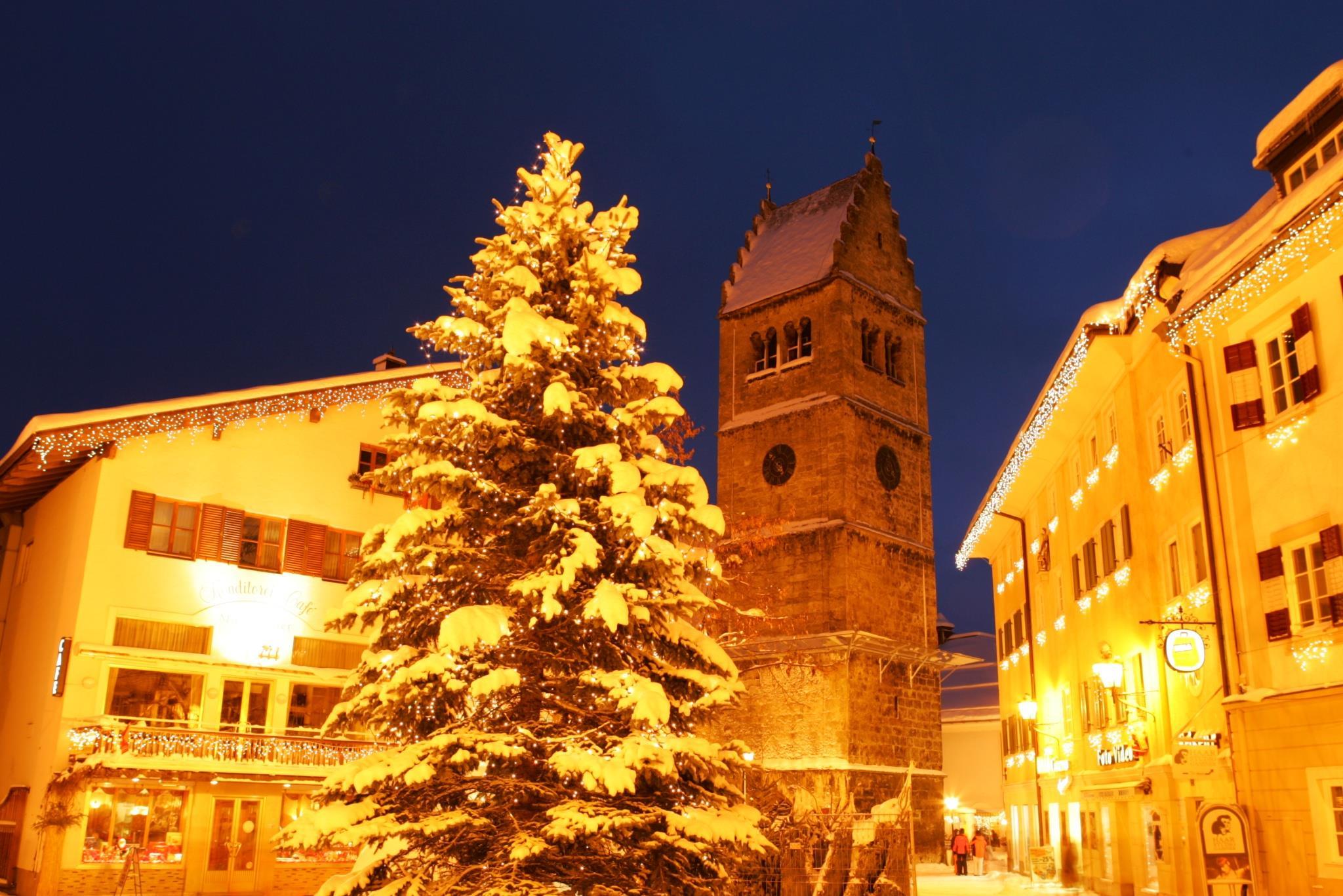 A snowy winter evening with a festively decorated Christmas tree. In the background, a historic tower and illuminated buildings can be seen.