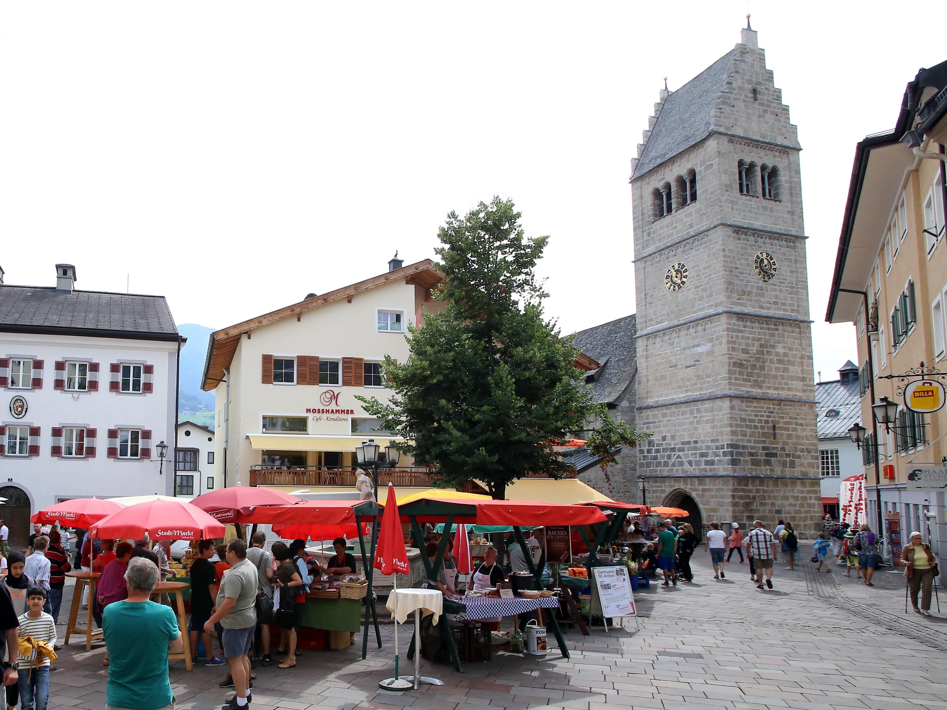Ein belebter Marktplatz mit bunten Ständen und Menschen, die einkaufen. Im Hintergrund steht ein historischer Glockenturm.