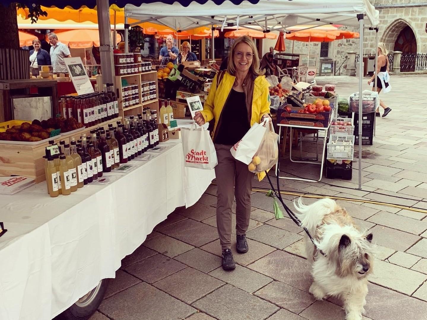Eine Frau mit einem Hund besucht einen Markt. Sie trägt eine gelbe Jacke und hält Tüten mit Lebensmitteln in der Hand.