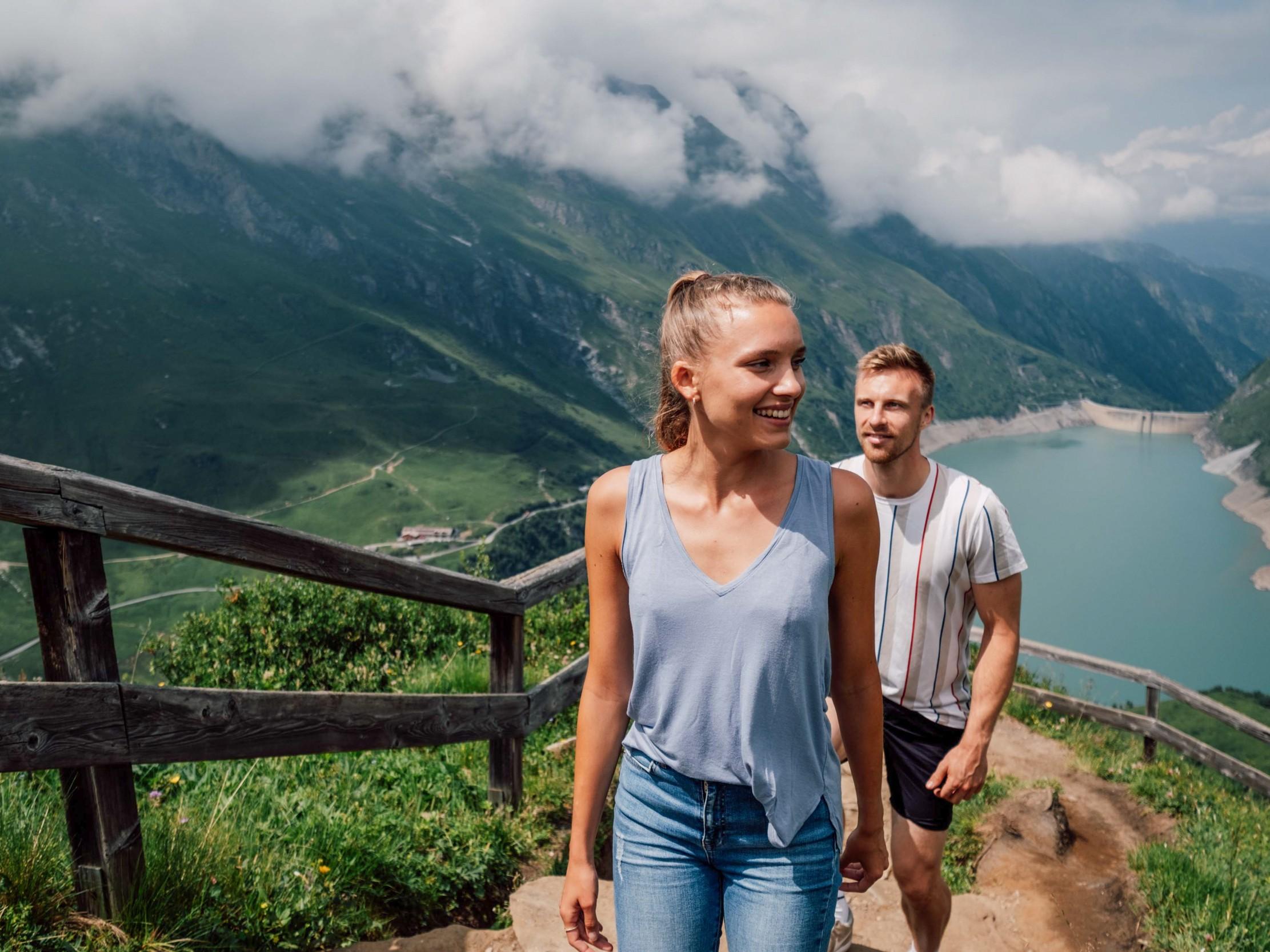 Ein Paar wandert auf einem schmalen Weg mit Blick auf einen Stausee und die umliegenden Berge. Der Himmel ist teilweise bewölkt und die Landschaft ist üppig grün.