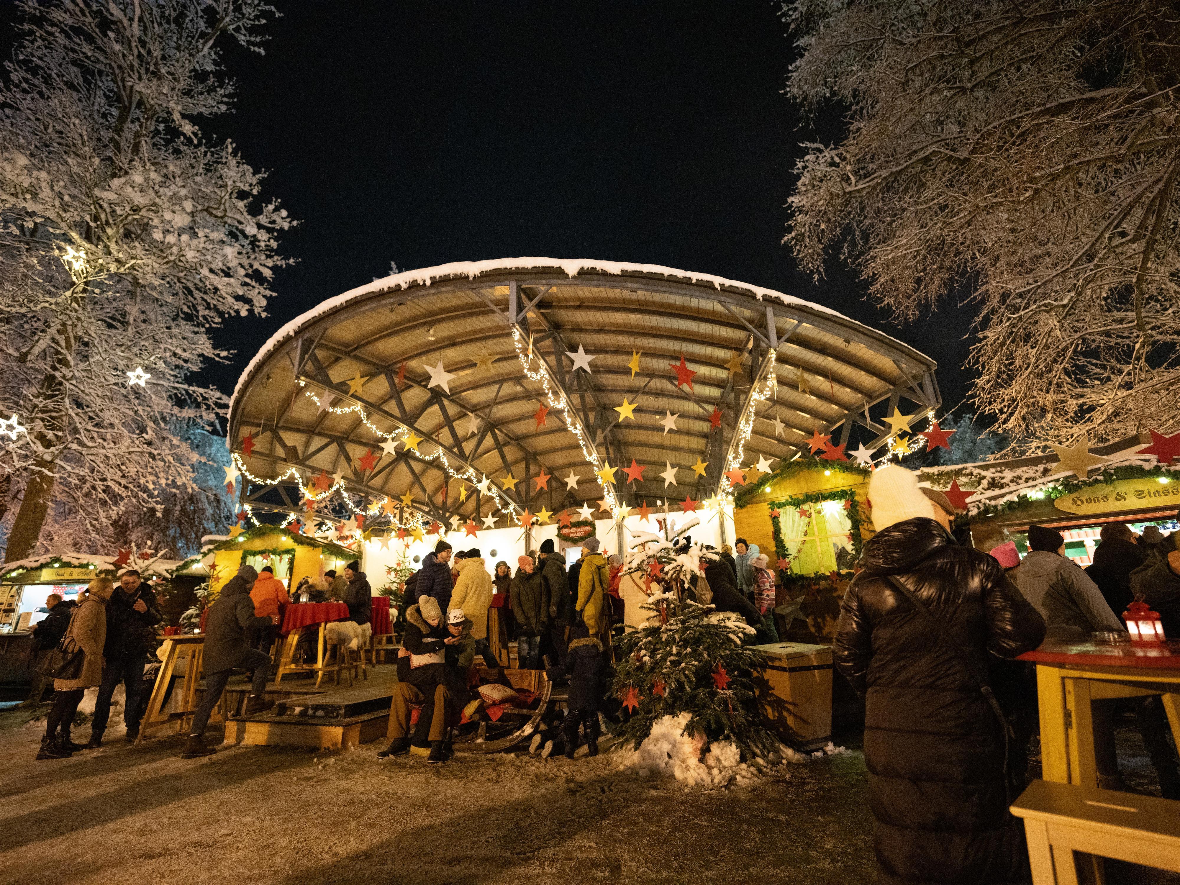 A winter Christmas market with festively lit stands. The surroundings are covered in snow and the trees shine in the warm light.