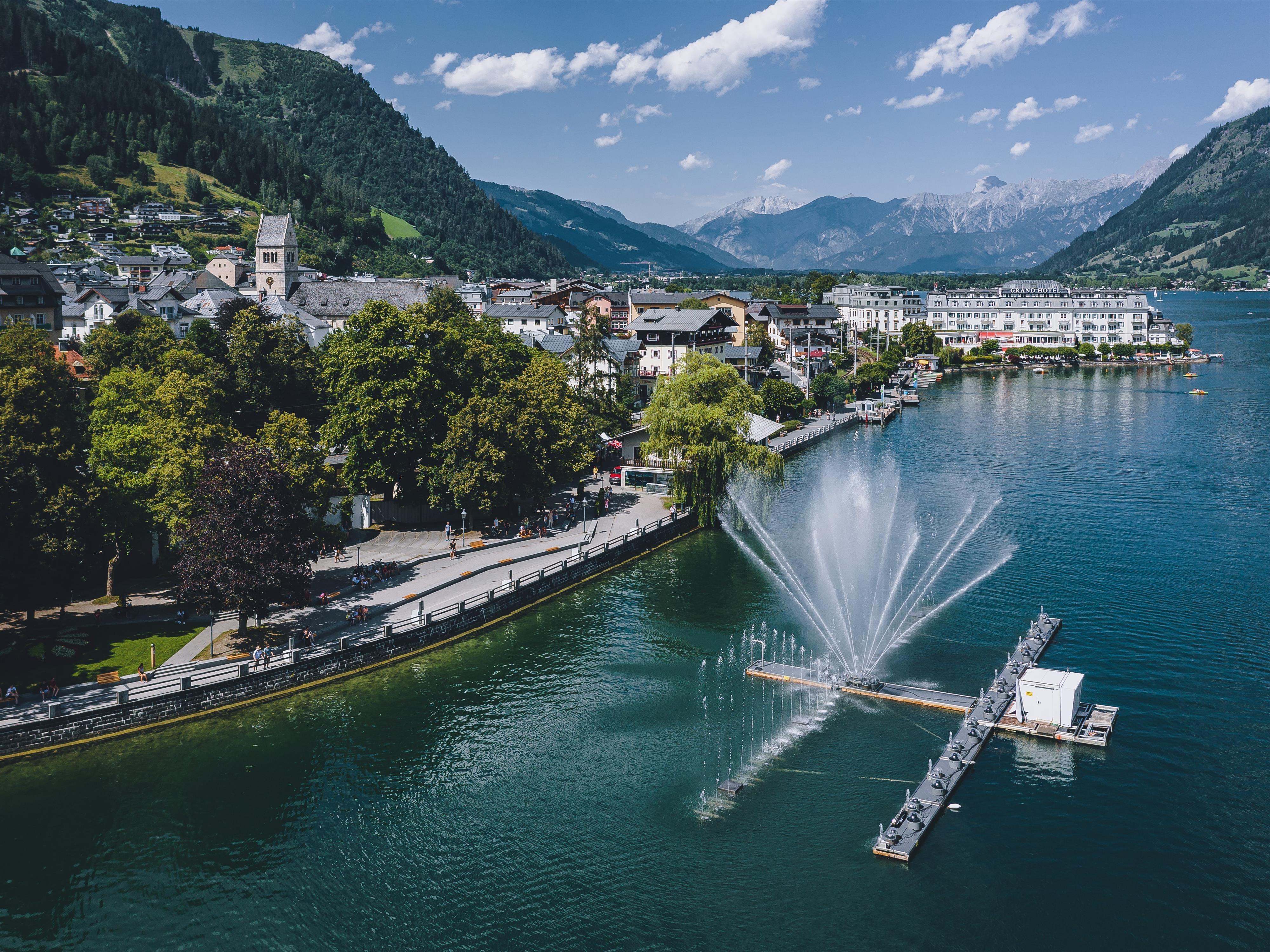 Eine malerische Landschaft mit einem klaren See und Bergen im Hintergrund. Im Vordergrund spritzt ein großer Brunnen Wasser in die Luft.