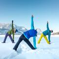 Drei Personen in warmer Sportbekleidung dehnen sich im Schnee. Im Hintergrund sind schneebedeckte Berge und ein blauer Himmel zu sehen.