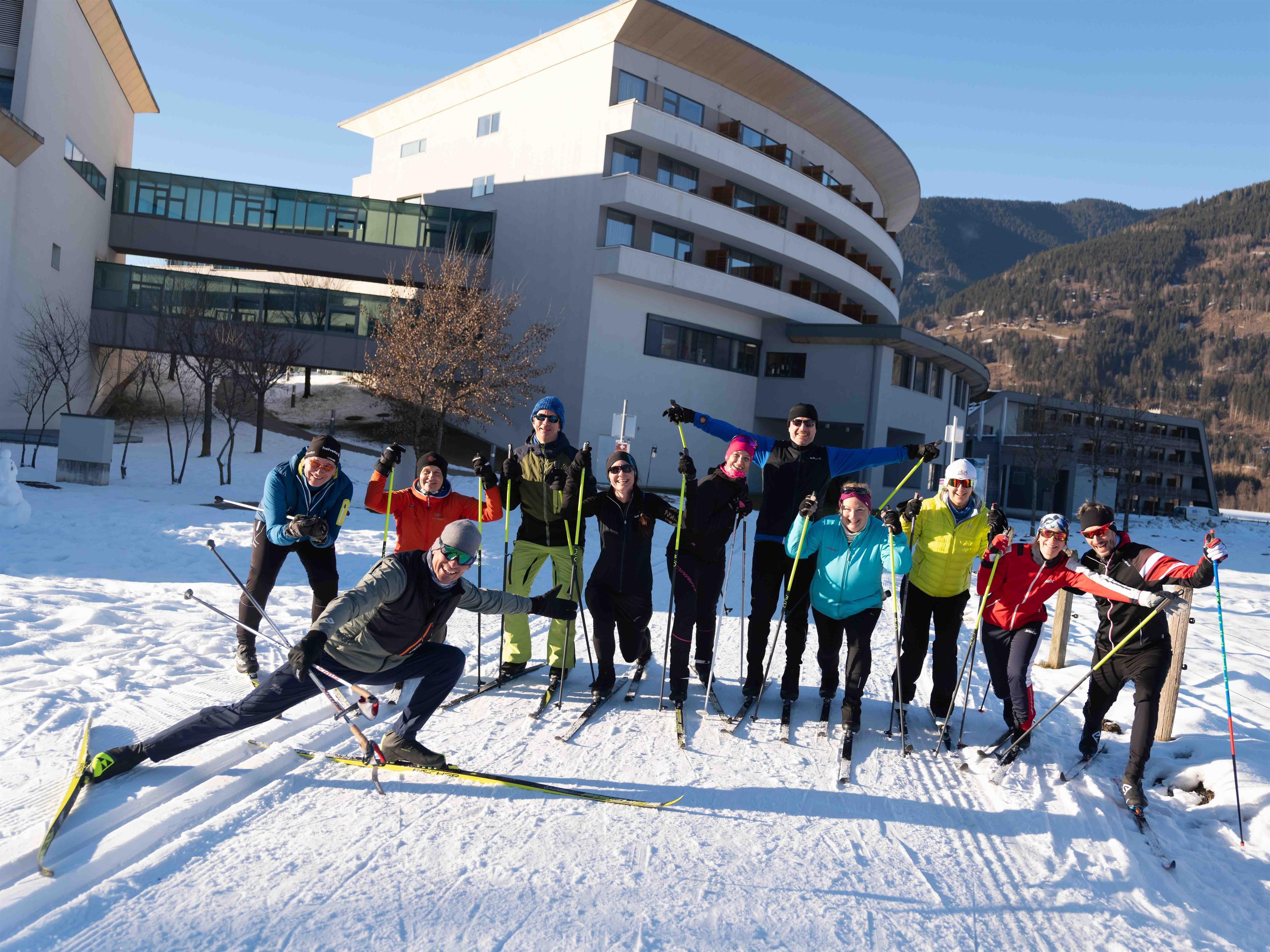 Eine Gruppe von Personen steht fröhlich im Schnee in Skiausrüstung. Im Hintergrund sind moderne Gebäude und Berge zu sehen.