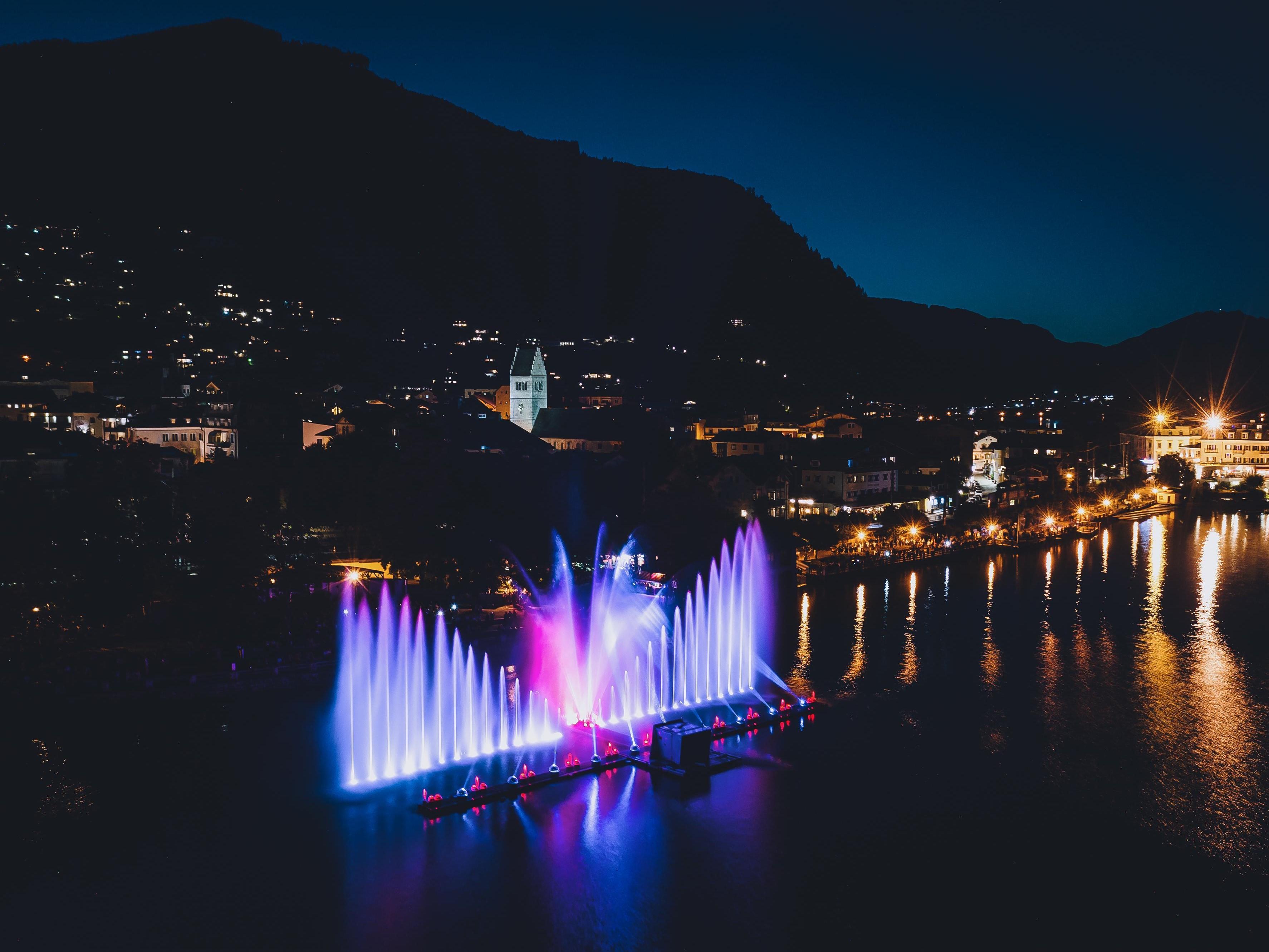 Eine lebendige Wasserfontäne mit bunten Lichtern beleuchtet den Abendhimmel. Im Hintergrund sind sanfte Hügel und Stadtlichter zu sehen.