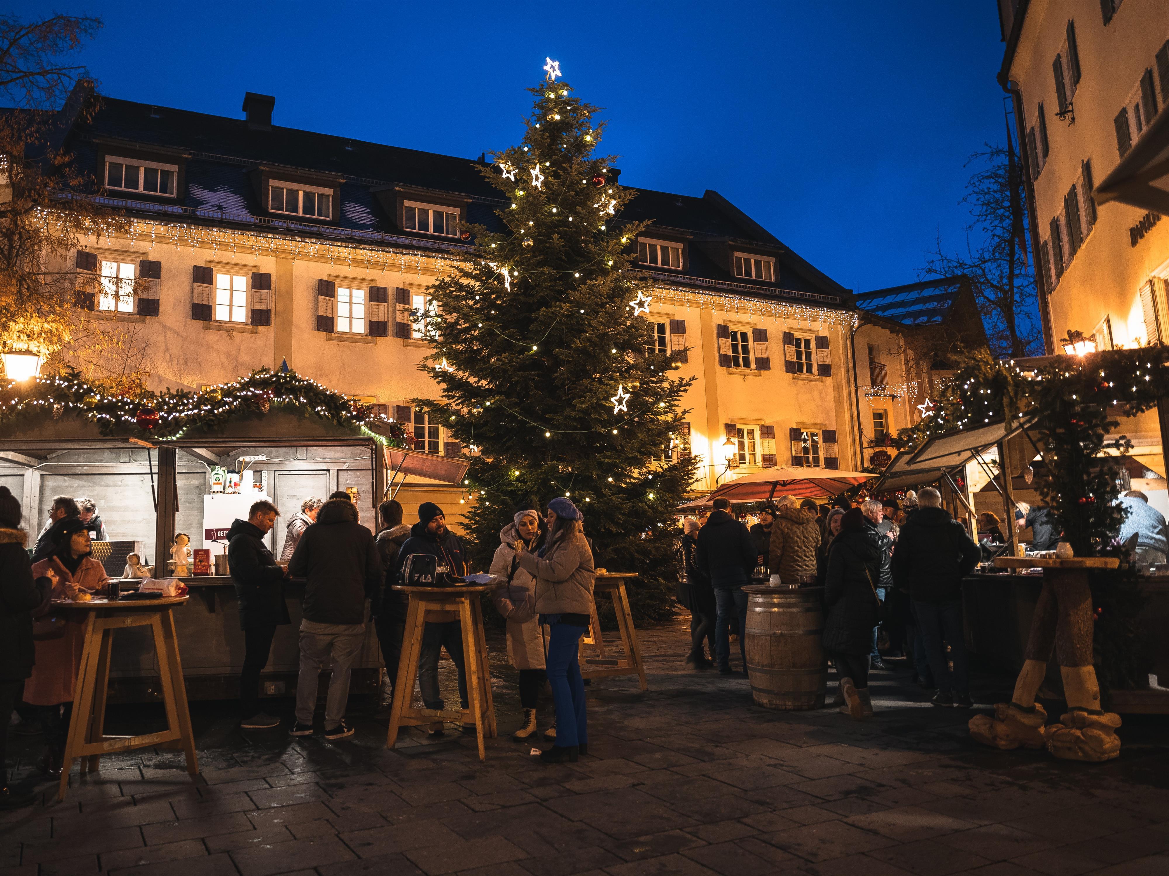 Ein festlicher Weihnachtsmarkt bei Nacht mit einem beleuchteten Tannenbaum. Menschen stehen an Ständen, genießen Getränke und Speisen in einer gemütlichen Atmosphäre.